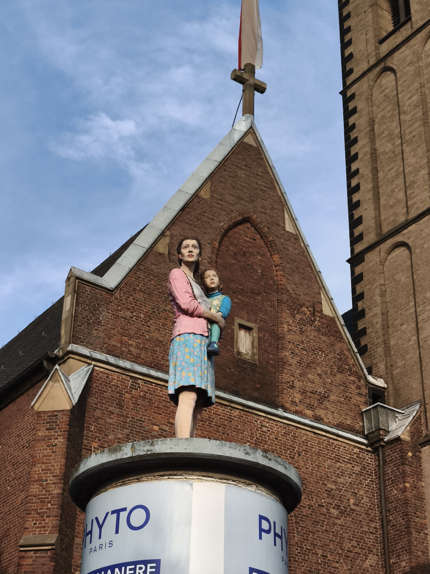 The image depicts a statue of a woman holding a child, positioned atop a cylindrical base on the facade of a brick building. The building appears to be a church, evidenced by a cross and a section of a tower visible. The statue is life-sized or larger and painted in vibrant colors. The woman wears a pink top and a blue skirt with a floral pattern, while the child is dressed in a blue outfit. The cylindrical base displays lettering, including "PHYTO PARIS" and "MANERE," suggesting it is part of an advertisement or monument. The statue is viewed from below, with a blue sky and white clouds in the background. This monument is located at the **Jan-Wellem-Denkmal** in **Dusseldorf, Germany**, specifically on the **Ritterstraße**.