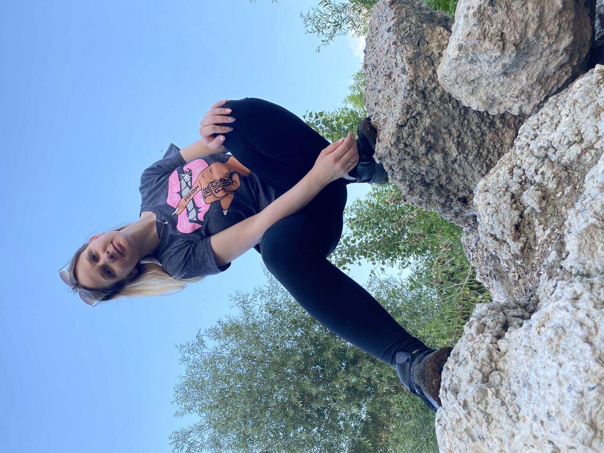 A woman with blonde hair is posing for a photo outdoors. She is wearing a dark t-shirt with a graphic print and black leggings. Her sunglasses are perched on her head. The woman is sitting with her legs crossed, and her hands are resting on her knees. She is looking directly at the camera with a neutral expression.
The background consists of a clear blue sky, lush green trees, and large rocks. The rocks appear to be rough and textured, possibly part of a natural formation or man-made structure. The lighting suggests it is daytime, likely a sunny afternoon. The overall scene conveys a relaxed, casual atmosphere.
The graphic on the woman's t-shirt includes text that reads "THE FUCK UP." This is the only visible text in the image. The woman is positioned in a way that makes her appear to be looking up from a lower vantage point, with the sky and trees creating a natural backdrop. The image does not provide specific clues about the exact location, other than the general context of being outdoors in a natural or semi-natural setting.