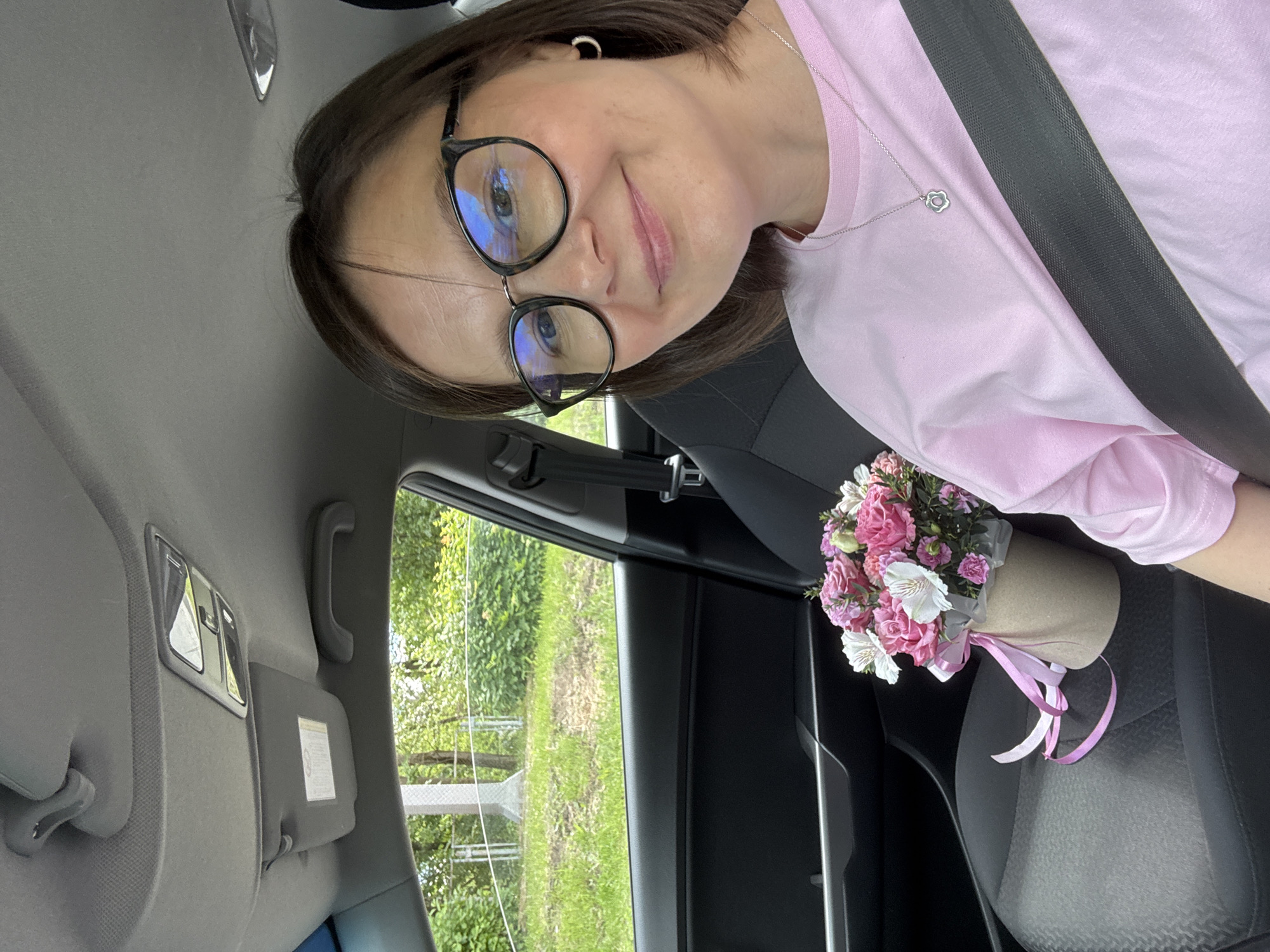 A woman with short brown hair and glasses smiles into the camera while sitting in the driver's seat of a car. She is wearing a pink t-shirt and a silver pendant necklace. A seatbelt is buckled across her chest. In the passenger seat, a bouquet of pink and white flowers in a cardboard box with a pink ribbon is visible. Outside the car window, green trees and grass can be seen. The car interior features a gray headliner, sun visor, and black car seats. The woman is holding a bouquet of flowers, suggesting a gift or celebratory occasion.