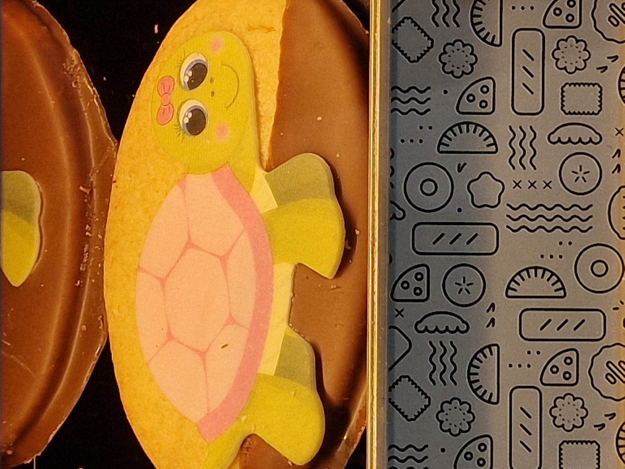 The image depicts two iced cookies and a patterned container. On the left is a brown, oval-shaped cookie with a yellow central area. Adjacent to it is a yellow oval cookie decorated with a pink and green cartoon turtle. The container on the right has a blue-grey background with black line drawings of food items. The location, Durham, United Kingdom, cannot be determined from the image.