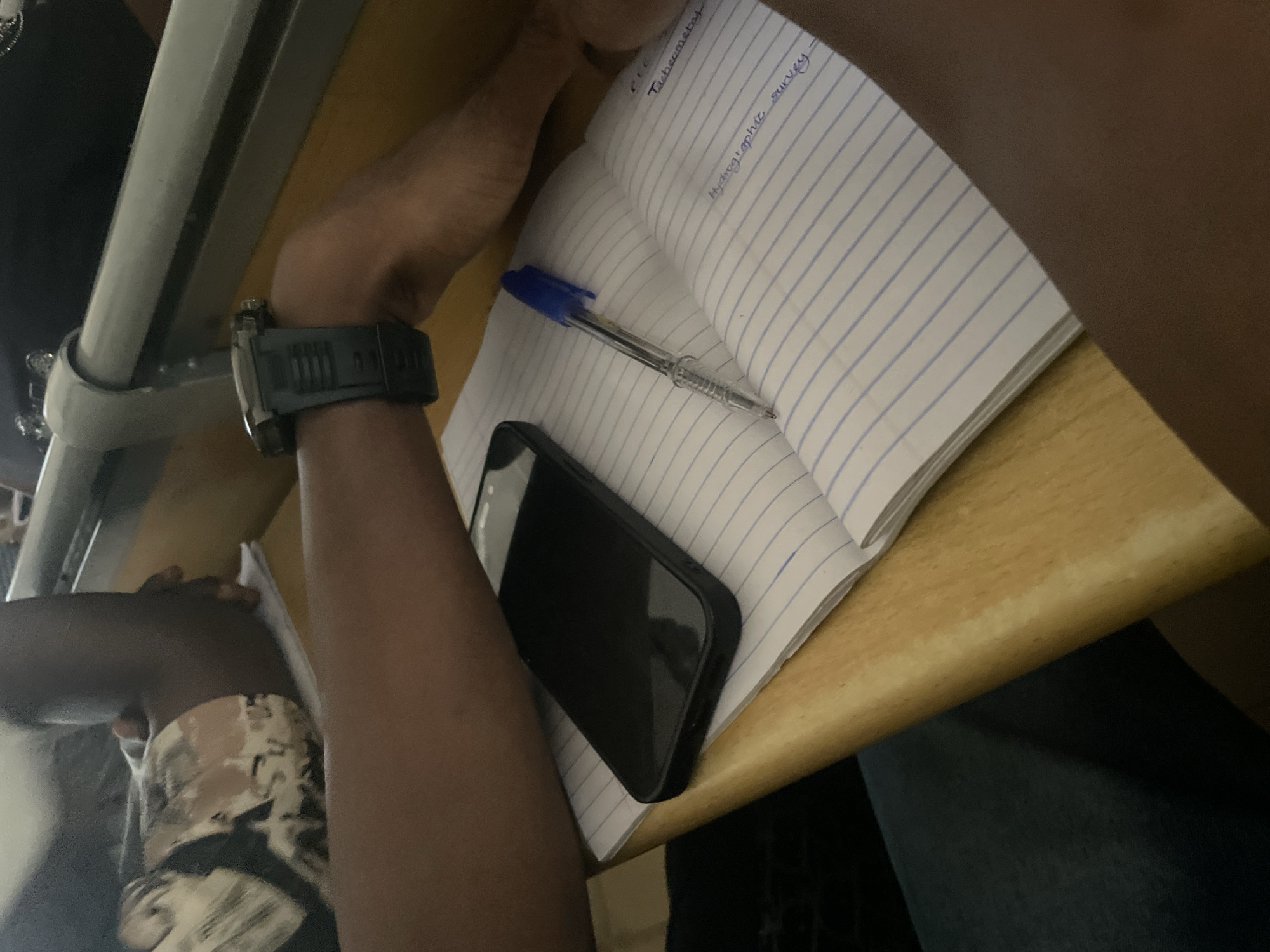 This is a close-up, eye-level, indoor shot of a person's arms and a notebook on a wooden desk. The notebook is open to a page with blue ruled lines. Handwritten text is visible on the right page, including "Tachemetes" and "Hydrographic survey." A black smartphone is lying face down on the left page of the notebook. A blue ink pen rests on the open pages. The person is wearing a dark-colored watch on their left wrist. The background is blurred, showing hints of a person in a patterned shirt and a grey metallic rail. The lighting suggests an indoor setting, possibly a classroom or study area. The provided crops offer closer views of the person's watch, the phone and notebook, and the handwritten text.
While the image depicts elements commonly found in a city, the specific location of Ihiagwa, Nigeria, cannot be determined from the visual information presented in the image.