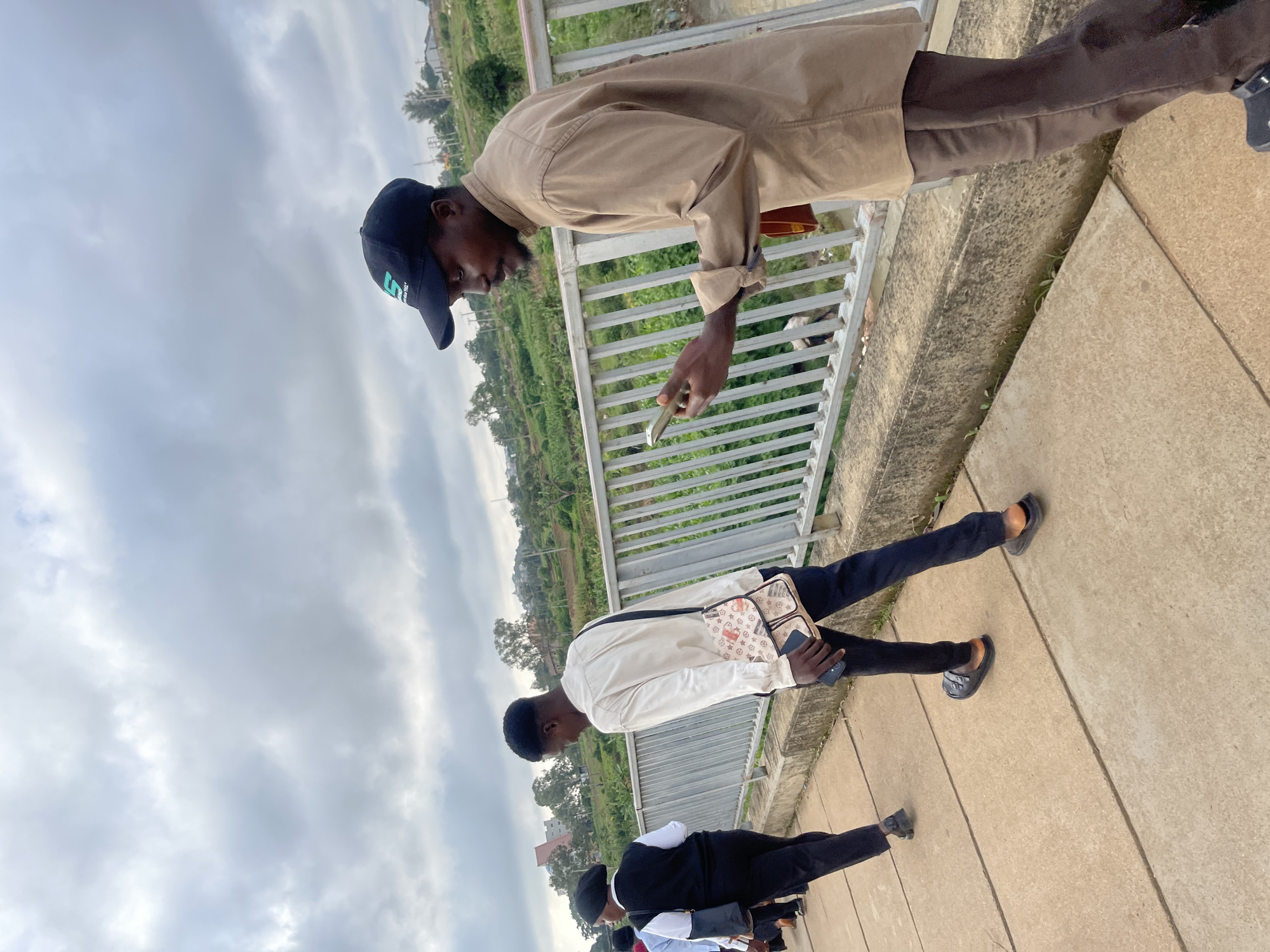 This image captures an outdoor scene with overcast skies. In the foreground, a person wearing a brown long-sleeved shirt, dark pants, and a black baseball cap is walking and looking at their phone. They are holding a brown object in their right hand. To their left, another individual in a white long-sleeved shirt, black pants, and a black vest over the shirt is walking away from the camera. They are carrying a patterned crossbody bag. Further behind them, other people are partially visible walking along a paved walkway. A metal railing runs along the right side of the walkway, separating it from a vegetated area and a body of water or a steep drop. The environment suggests an urban or semi-urban setting, possibly a bridge or elevated walkway, given the railing and the view of buildings and greenery in the background. The location is identified as Rafin Par, Nigeria.