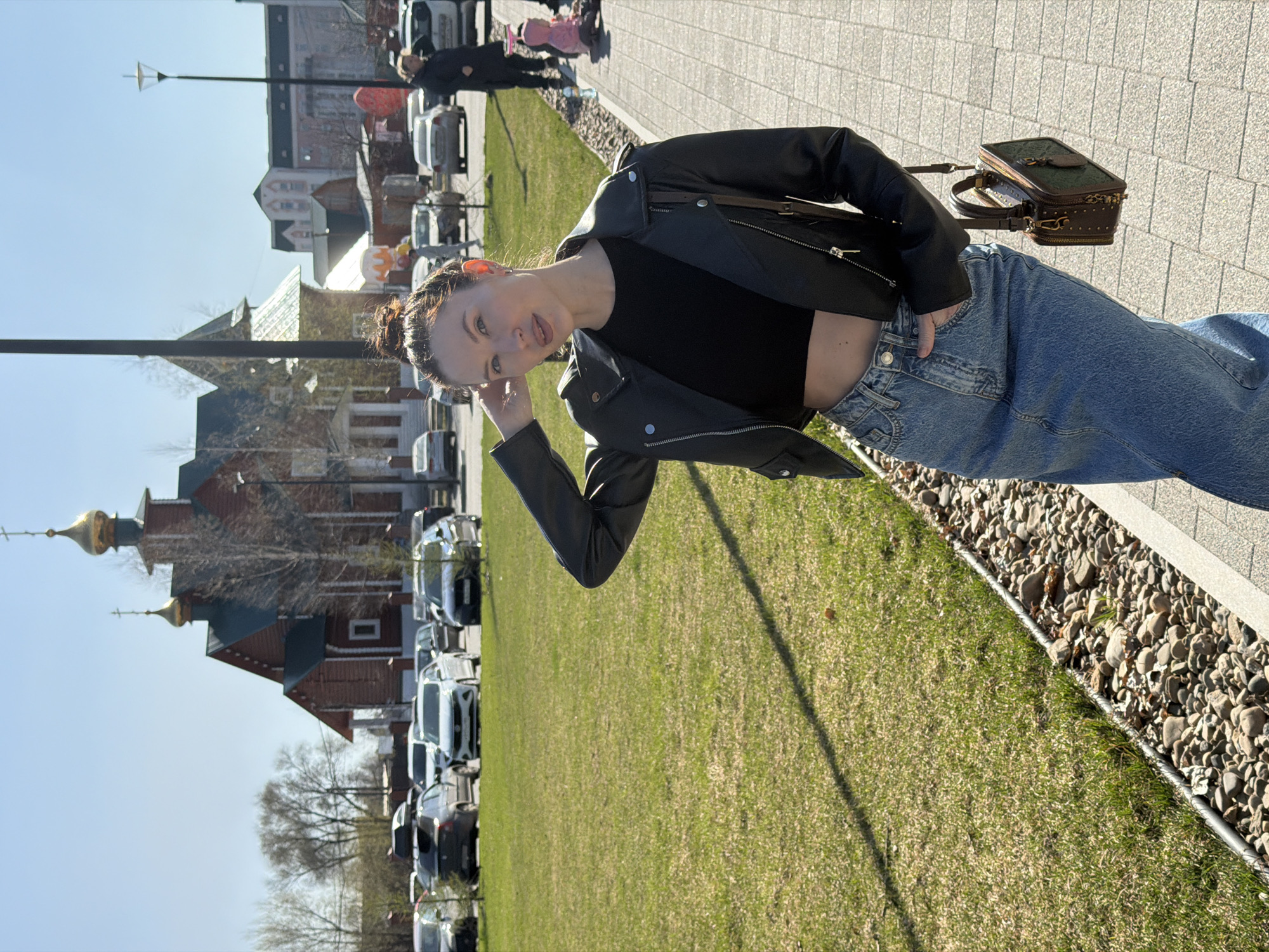A young woman is photographed in Perm, Russia, likely near the Holy Trinity Stefan Monastery, indicated by the distinct golden domes of the church structure visible in the background. She stands on a brick-paved path bordered by a grassy area with a line of parked cars and other buildings further behind. The woman is wearing a black crop top, a black leather jacket, and blue jeans. She carries a brown and green patterned bag over her shoulder. Her hair is up, and she is looking at the camera, holding one hand up near her head. Other people, including someone pushing a stroller, are further back along the path. The sky is clear and blue.