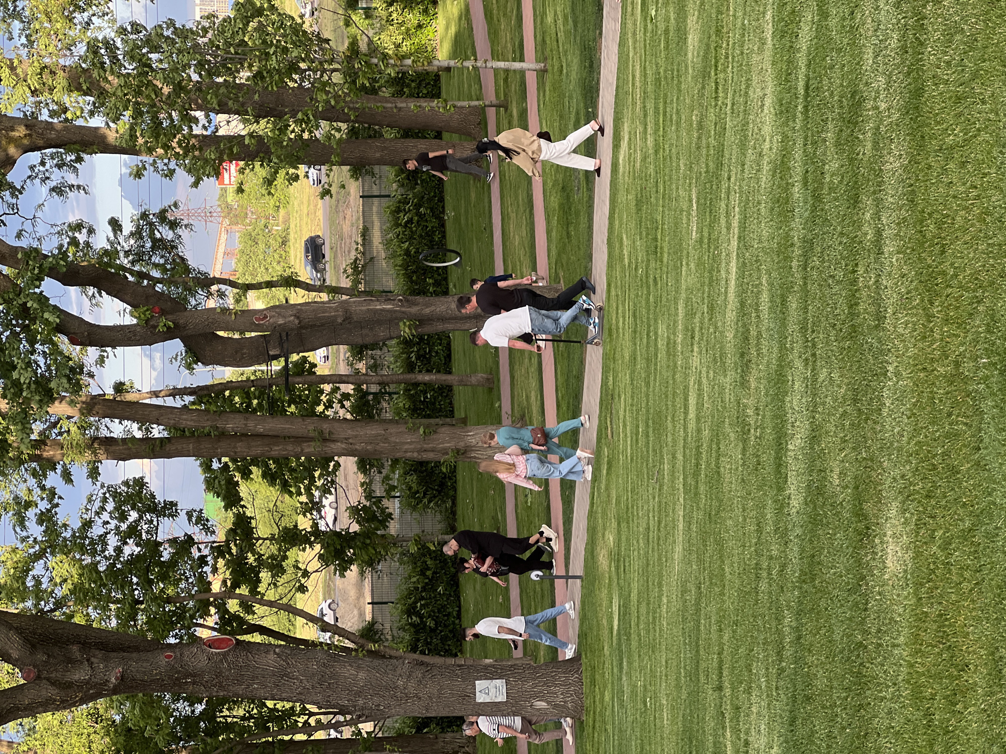 The image depicts a park setting. The grass is a vibrant green and appears to be neatly manicured. Trees line the edge of the grassy area, providing shade and creating a natural boundary. A group of people is present. Some are walking or strolling along the paved path, while others appear to be engaged in activities such as riding a scooter. The environment suggests a public park or recreational area, possibly located in Krasnodar, Russia. In the background, a crane construction and buildings are visible indicating an urban area.