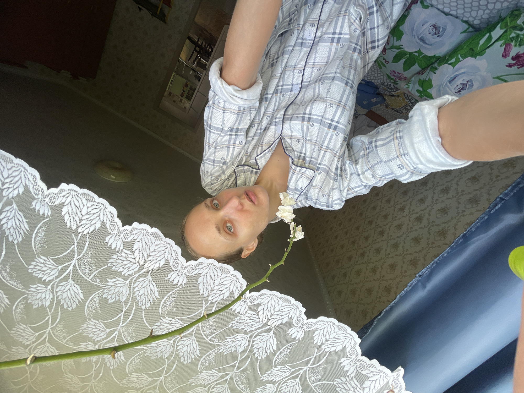 This is a vertical, slightly overhead, close-up selfie taken by a woman indoors. The woman, with light skin and blue eyes, is wearing plaid pajamas and looking directly at the camera with a neutral expression. A thin stem with a few small white flowers extends from the lower left of the frame towards her face. A white lace tablecloth or curtain with a leaf pattern is in the foreground, obscuring the bottom portion of the image. Behind the woman, a bed with floral patterned pillows is visible, featuring large blue roses. The room appears to be dimly lit, suggesting it might be evening or daytime with closed curtains. The wallpaper in the background has a repeating floral motif. The image is rotated 90 degrees counter-clockwise.