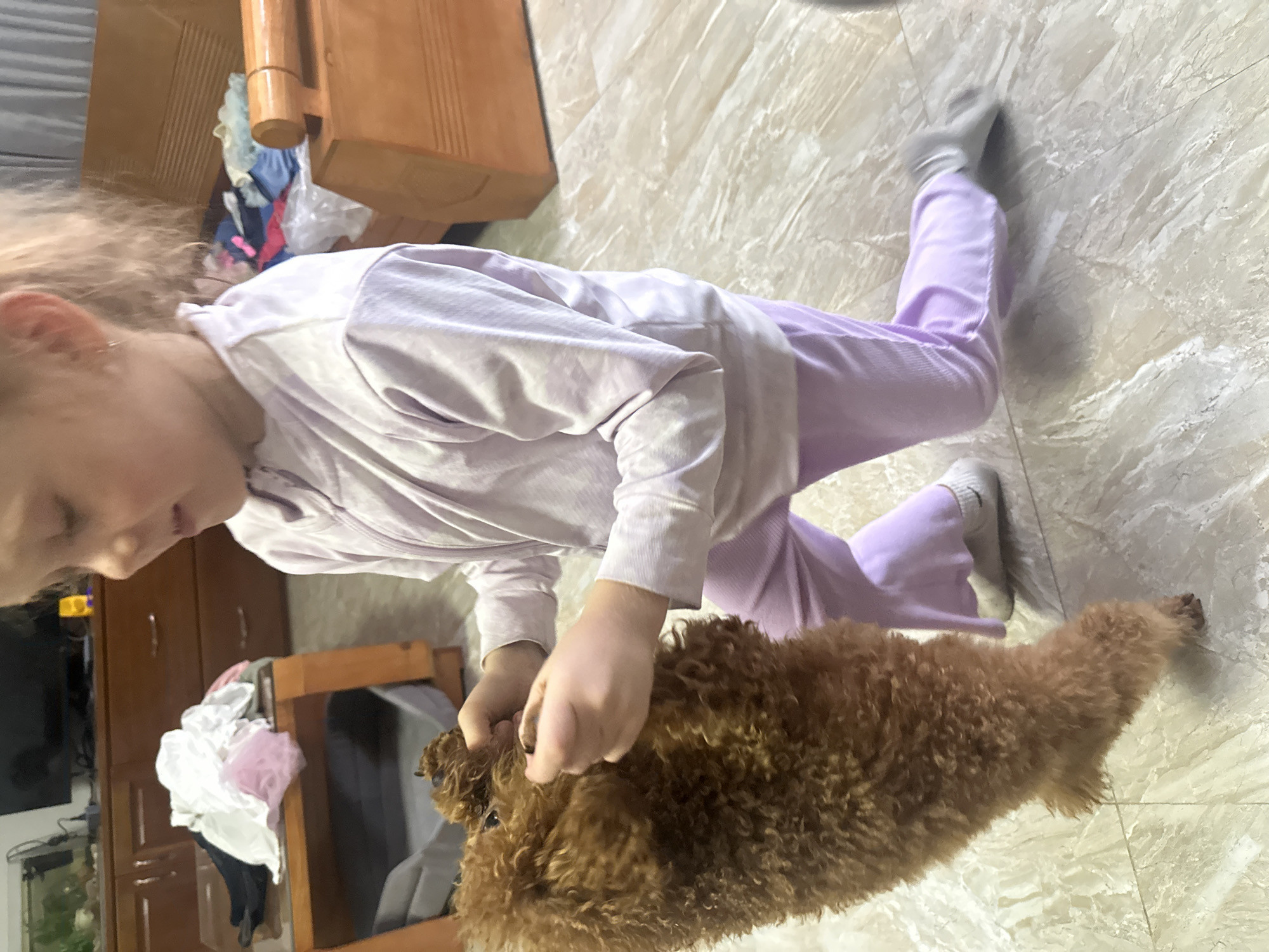 A child, likely a girl due to her blonde hair and soft-colored pajamas, is on the floor interacting with a small brown poodle dog. The child wears a light purple long-sleeved shirt and light purple pants, with gray socks on her feet. She is crouched down and appears to be petting the dog. The floor is tiled with a light beige, marbled pattern. In the background, there is furniture that appears to be made of wood, including a cabinet with clothes piled on top. A possible fish tank and other belongings are also visible. The scene appears to be indoors, possibly a home setting.
Based on the city name, it seems likely this location is in Bac Nha Trang Ward, Vietnam.