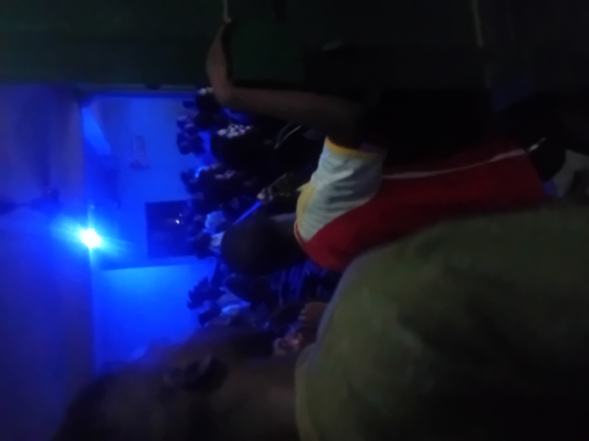 A dimly lit, rotated image appears to depict a person in a red and white shirt in an indoor setting. Behind the person, there is what appears to be a wall illuminated with blue light. The context of the location and activity is not discernible due to the poor lighting and limited visible details.