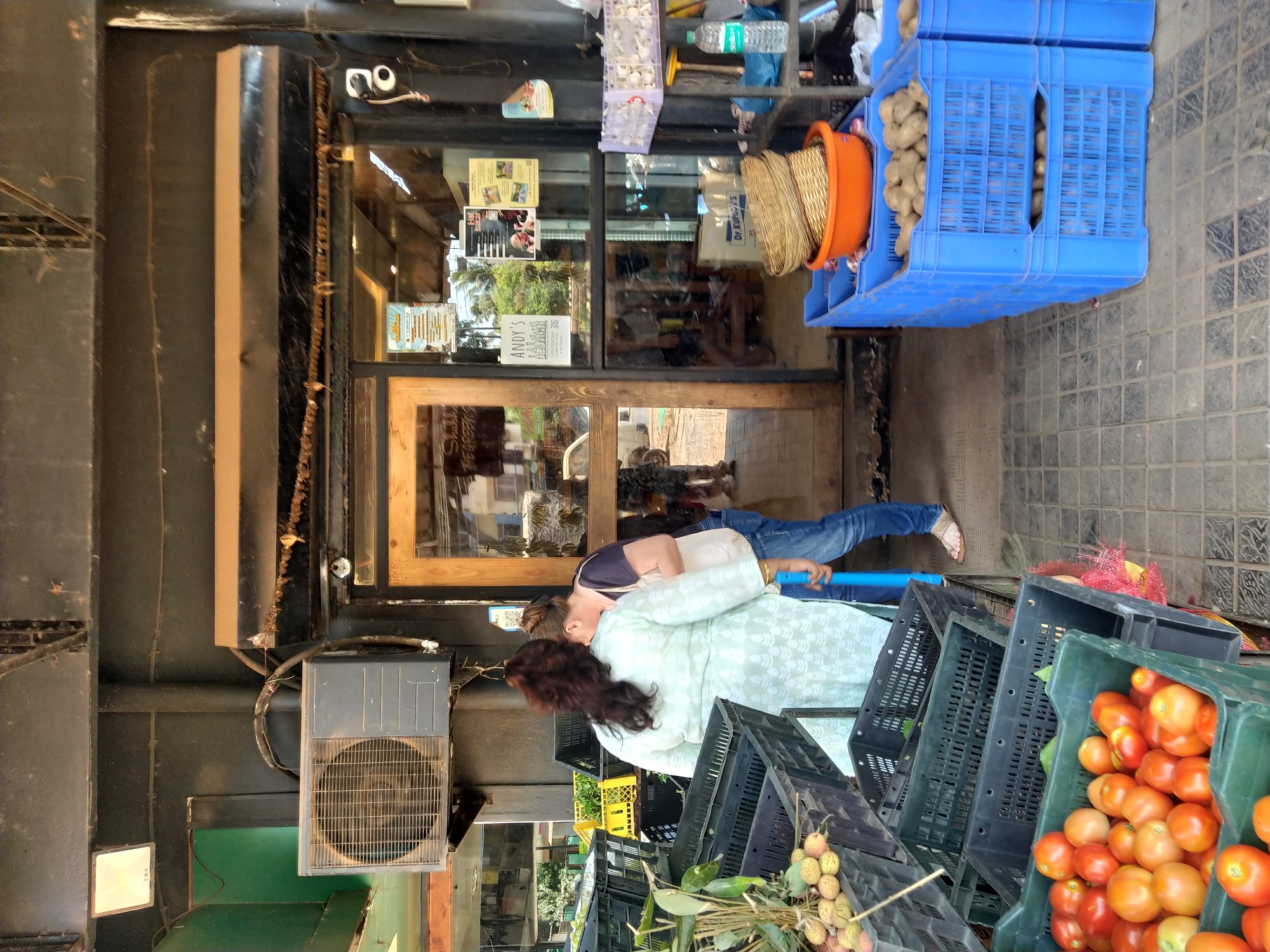 The image depicts a street scene in Oxel, India, in front of a store named Andy's. Two individuals are positioned outside the store. One person, wearing jeans and a blue shirt, is standing to the right of a second individual, who is wearing a light green dress. The second individual is positioned among several stacks of black and green plastic crates filled with produce, including tomatoes and lychees. To the right of the individuals are blue plastic crates, some containing potatoes and onions in an orange bucket. The store's entrance features a wooden-framed glass door with signs and images posted on the glass. An air conditioning unit is mounted on the exterior of the building to the left. The scene suggests an outdoor market setup in front of a small store.