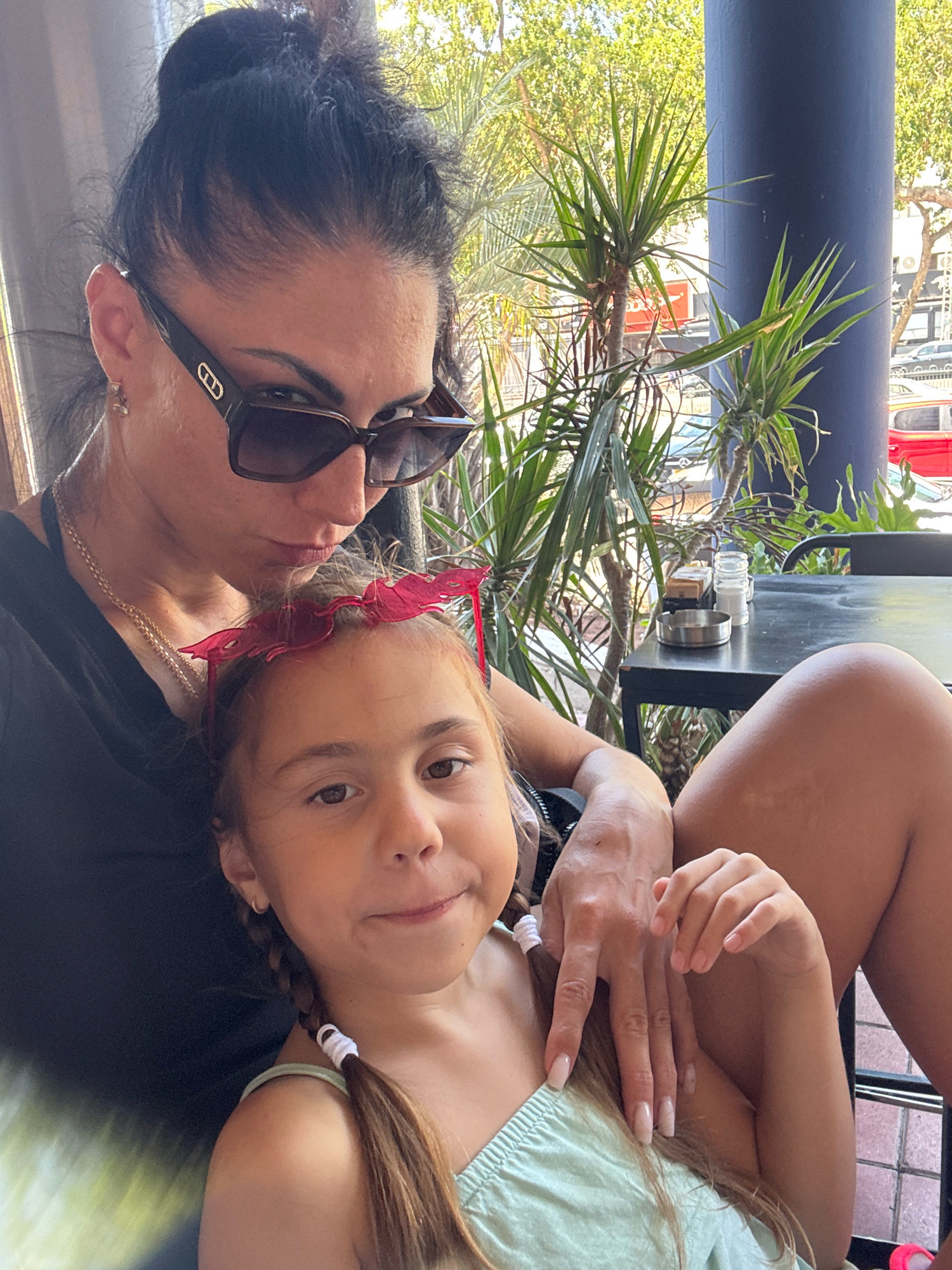 A woman and a young girl are posing for a selfie, outdoors at a cafe, possibly in Netanya, Israel, as indicated by the presence of Dracaena trees and Mediterranean architecture visible in the background. The woman, wearing a black top and large sunglasses, is holding the girl, who has pink novelty glasses on her head. The girl is wearing a light green top. The scene suggests a relaxed, casual atmosphere, with trees and a dark blue column forming the backdrop.
