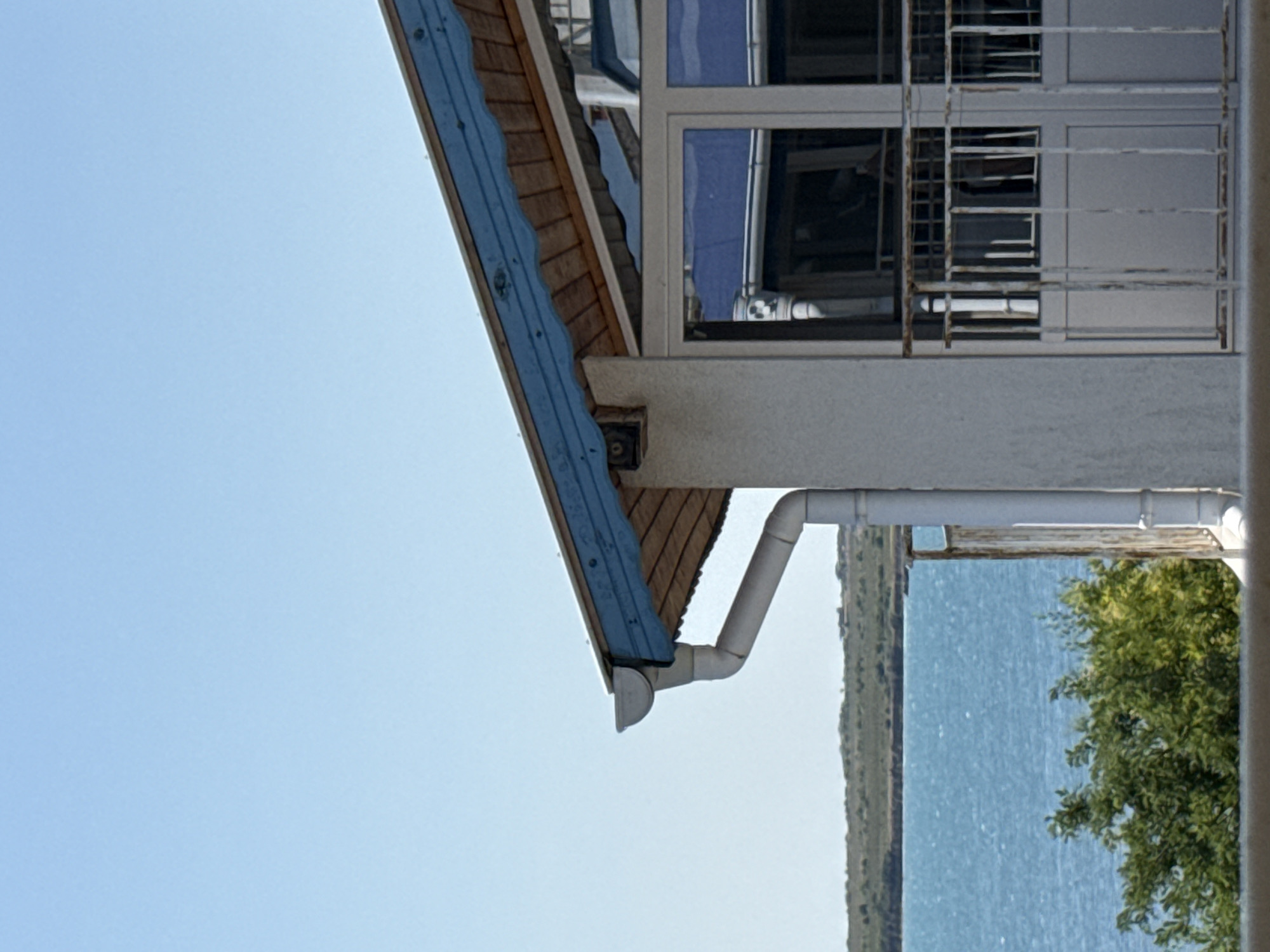 A close-up, eye-level shot captures a portion of a building's exterior with a scenic backdrop. The building features a white stucco wall and a roof with a distinctive blue-painted wooden fascia, beneath which exposed wooden beams are visible. A white downspout system is attached to the side of the building. To the right, a balcony with a white railing and a metal grate overlooks the scene. Large windows with reflective surfaces show distorted reflections of the building's interior and exterior.
Beyond the building, a body of tranquil blue water stretches towards a distant, low-lying, green-covered coastline. The sky above is a clear, bright blue. Lush green foliage is visible in the foreground, partially obscuring the view of the water and coastline. The image appears to be taken on a clear, sunny day. The location is identified as the city of Pomorie, Bulgaria.