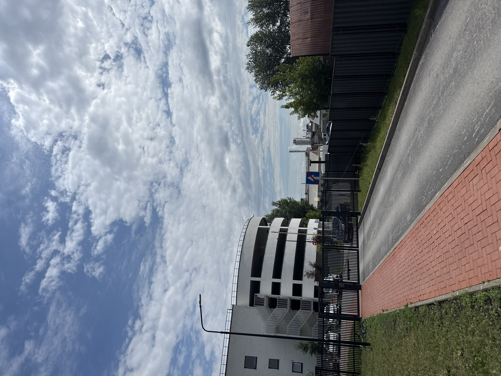 The image depicts an outdoor scene with a modern, multi-story white parking garage on the left. The garage has a curved facade with open levels, resembling a spiral. White outdoor stairs are attached to the side of the building. A tall black lamp post stands in front of the garage. A black metal gate encloses the entrance to a paved area. To the right of the gate, there's a red brick walkway. Beyond the gate, a road leads into the parking area where several dark-colored cars are visible. A dark metal fence runs along the right side of the road and walkway. In the background, there are trees, industrial buildings with smokestacks, and a cloudy blue sky. The scene appears to be in an urban or industrial area.