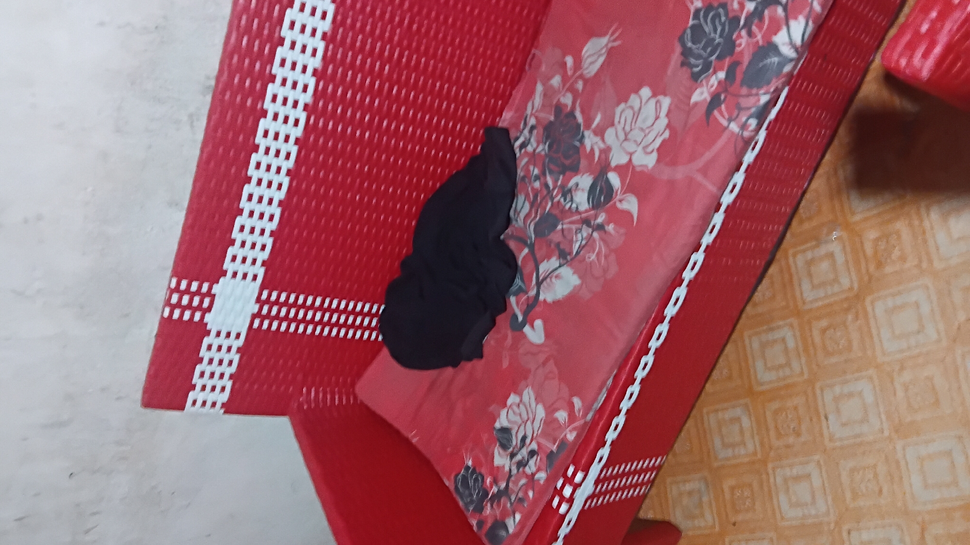 A red wicker sofa with a floral patterned mattress is shown in a room. A black head covering is draped on the mattress. The floor has a tiled pattern of squares within squares in a light orange color. The wall behind the sofa is a rough, textured off-white. The scene is located in Prabumulih, Indonesia.