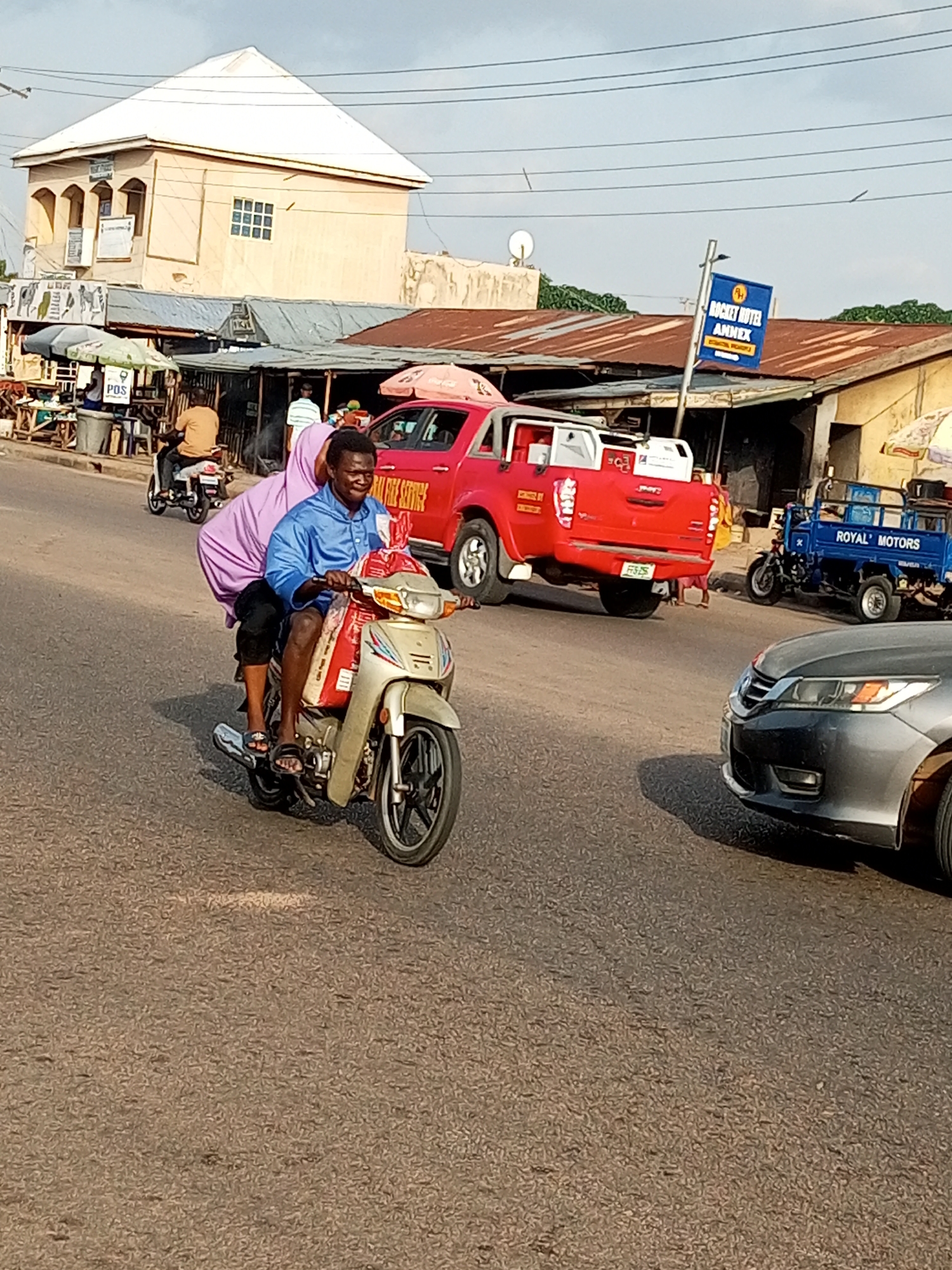 Two people are riding on a motorcycle. The person in front is driving, wearing a blue shirt. The person behind is wearing a purple garment and is holding onto the driver. They are both looking forward. The motorcycle appears to be silver and gold. In the background, a red pickup truck is visible. To the right of the truck, a blue three-wheeled vehicle with "ROYAL MOTORS" written on its side is parked. Behind the vehicles, there are buildings with corrugated metal roofs, suggesting a commercial area. A sign with "ROCKET HOTEL ANNEX" is also visible. The sky is overcast, indicating it might be daytime. The road surface is asphalt.