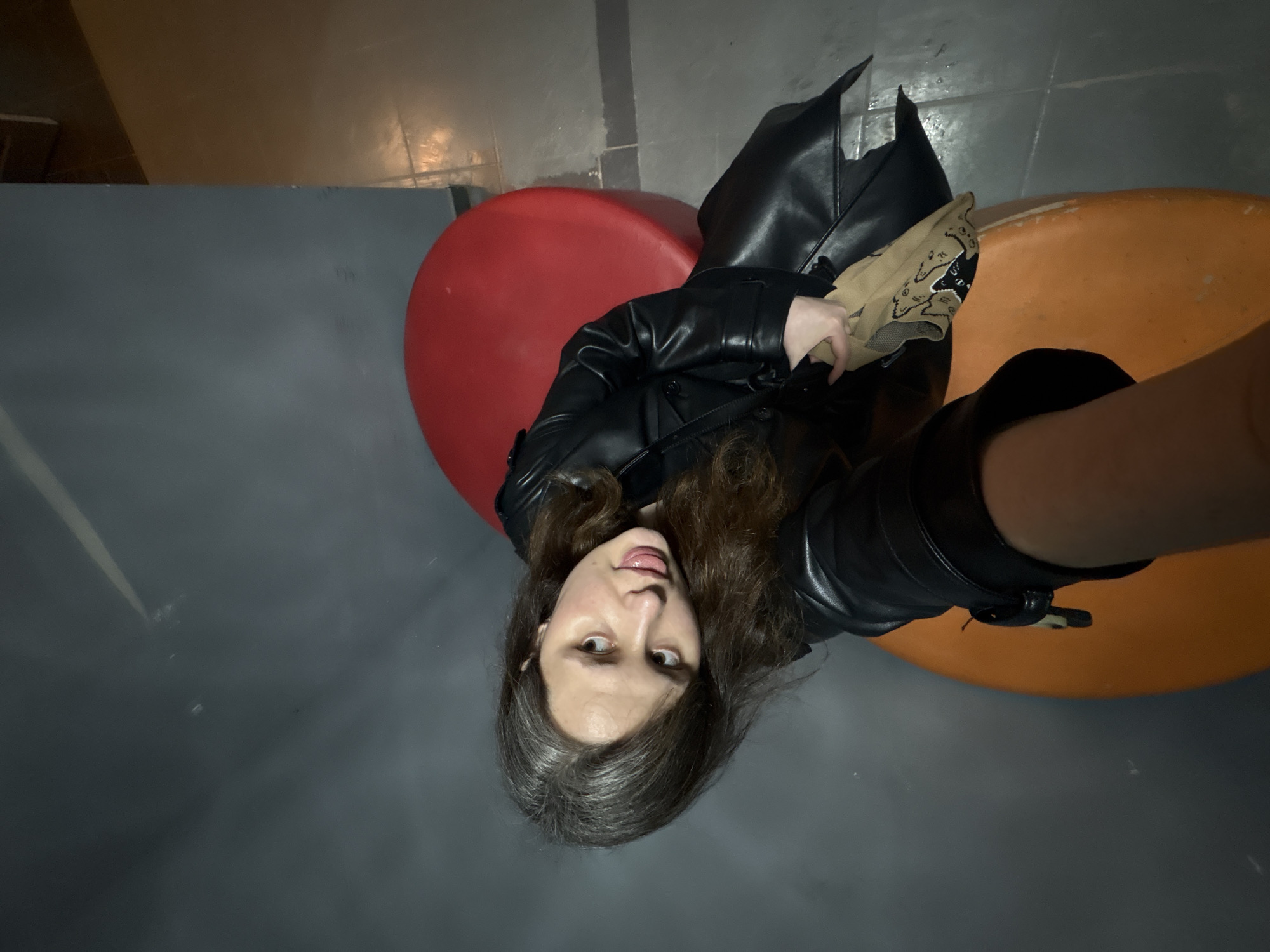 A high-angle, slightly off-center image depicts a female subject indoors. She has long, dark hair and is wearing a black leather trench coat. Her head is tilted, and she is looking directly at the camera with her tongue slightly extended. Her left hand is visible, holding a tan fabric item with black patterns. She is positioned between two large, rounded, cushioned objects resembling stools, one red and the other orange. The background consists of a plain grey wall and a tiled floor, which transitions from a dark grey in the foreground to a lighter brown or beige hue further back. The specific location within the city of Chaikovsky, Russia, cannot be determined from the visual information provided.