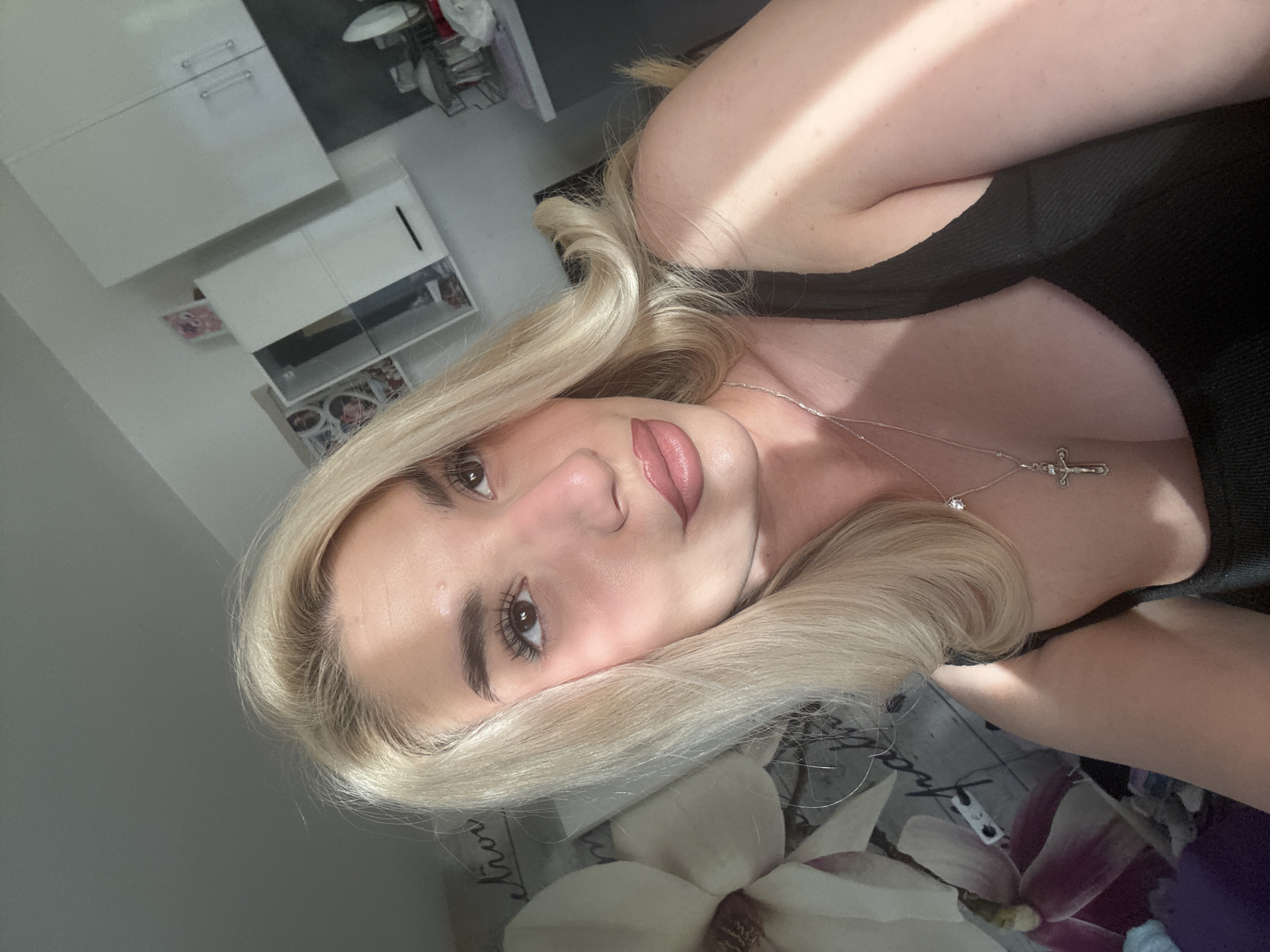 A light-skinned woman with blonde hair is depicted in an eye-level selfie, turned counter-clockwise. She has brown eyes and pink lips. She is wearing a black tank top and a silver necklace with a cross pendant. The background appears to be a residential space. Visible in the background is a white cabinet and a glimpse of what appears to be a kitchen rack. A printed floral design is partially visible under the woman. The location of this photo is unknown and there is no explicit data indicating that it was taken in Sofia, Bulgaria.