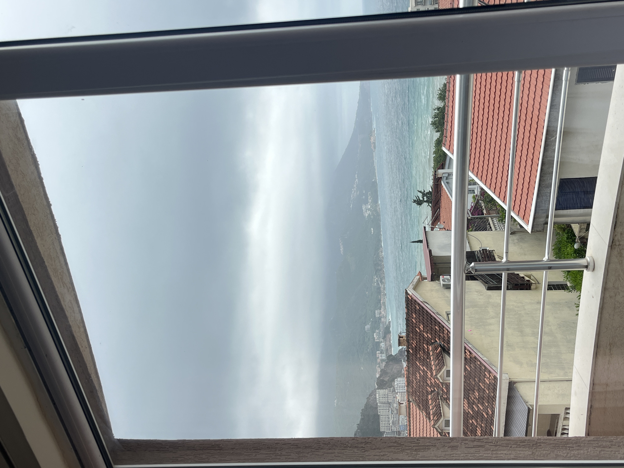 The image shows a view of the Adriatic Sea and the coastal town of Bečići, Montenegro, seen from what appears to be an elevated indoor location, possibly a balcony or a window. The sky is overcast, suggesting a cloudy or rainy day. In the foreground, elements of the building from which the photo is taken are visible, including part of the window or balcony structure. Neighboring buildings with red-tiled roofs are also in view. The sea has a somewhat rough surface, indicative of wind. The cityscape is partially obscured by mist or fog, which could be due to the weather conditions. The buildings seem to be clustered along the coastline and possibly extend up the slope of a hill.