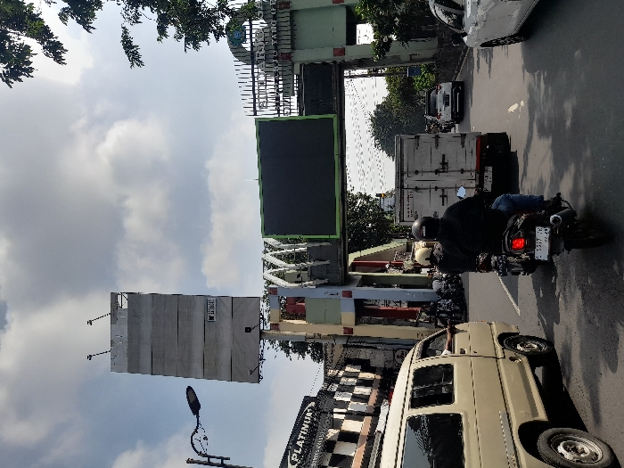 The image shows a street scene in Kecamatan Waru, Indonesia (latitude: -7.352, longitude: 112.729). A large billboard is located on the left side of the street, while a smaller billboard with a black screen is located on the right. A white van is driving on the left side of the street, and there are three motorbikes driving on the right side. The motorbikes are being driven by two males. One person is visible in the van. There are also a few cars driving in the distance. The environment is urban and the sky is partially cloudy. A street light is visible on the left side of the street.