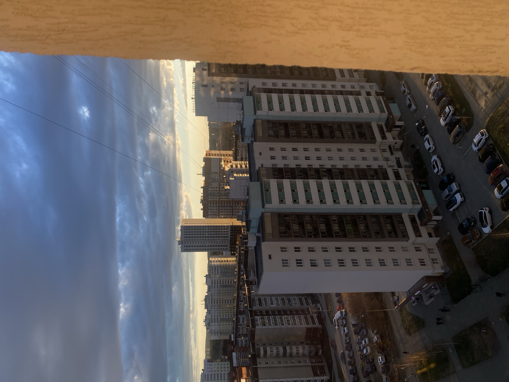 This image presents a high-angle urban landscape, likely captured from a tall building or balcony, overlooking a city at sunrise or sunset. The scene is dominated by numerous multi-story apartment buildings, stretching into the distance. Many buildings feature white and grey facades, with some showcasing light blue or green striped sections and multiple windows.
In the foreground, a light-brown, textured surface (possibly a balcony railing or wall) frames the top of the image. Below, a complex of modern residential buildings lines a street where a sizable parking lot is filled with various cars, including white, black, blue, red, and brown vehicles. Several streetlights are also visible.
The sky occupies a large portion of the frame, displaying a dramatic mix of clouds. Towards the horizon, clouds are illuminated with a golden glow from the low sun, creating a warm hue, while closer to the viewer, the clouds appear darker and more overcast. Several thin, dark power lines or cables horizontally traverse the sky.
On the ground level, near one of the buildings, small figures can be discerned, indicating people walking on sidewalks. One building's facade shows partially obscured Cyrillic text, possibly "СТРОЙ..." or "STROY...", suggesting a construction-related name or company.
The overall impression is one of a bustling city experiencing either the dawn or twilight hours, with the low sun casting long shadows and highlighting the architectural patterns of the urban environment, consistent with a setting like Yekaterinburg, Russia.