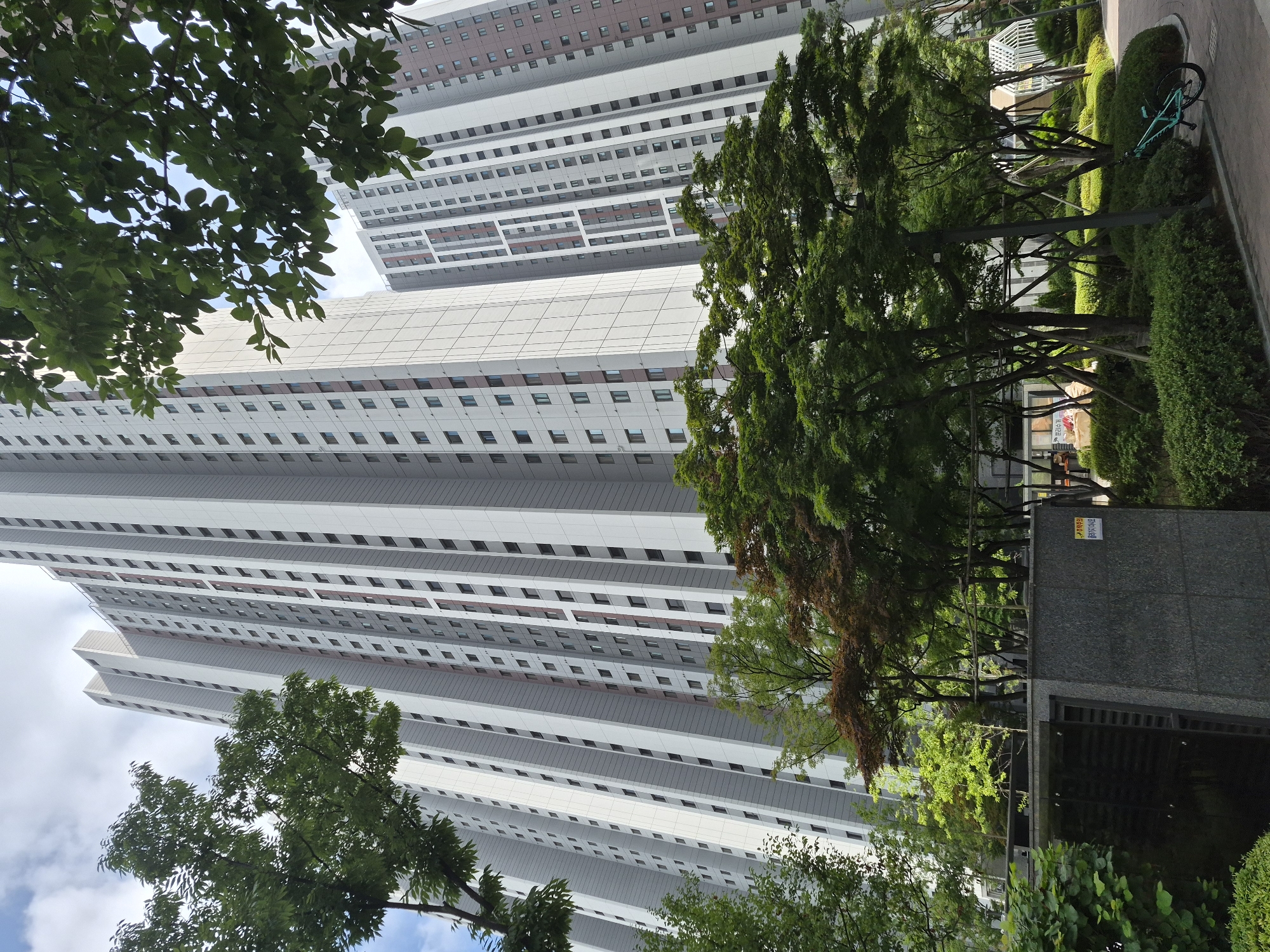 This is a photograph taken in the city of Ansan, South Korea, featuring a view of tall, modern apartment buildings. The buildings are primarily light gray and white, with darker accents around the windows and vertical design elements. Lush green trees and landscaping are prominent in the foreground and around the base of the buildings, suggesting a well-maintained urban environment. In the lower right of the image, a light blue bicycle is parked on a paved walkway, leaning against a low-lying shrub bed. A dark gray, cube-shaped structure, possibly an entrance or utility building, is visible in the lower left quadrant of the image. The sky is partially cloudy, indicating daytime conditions.
