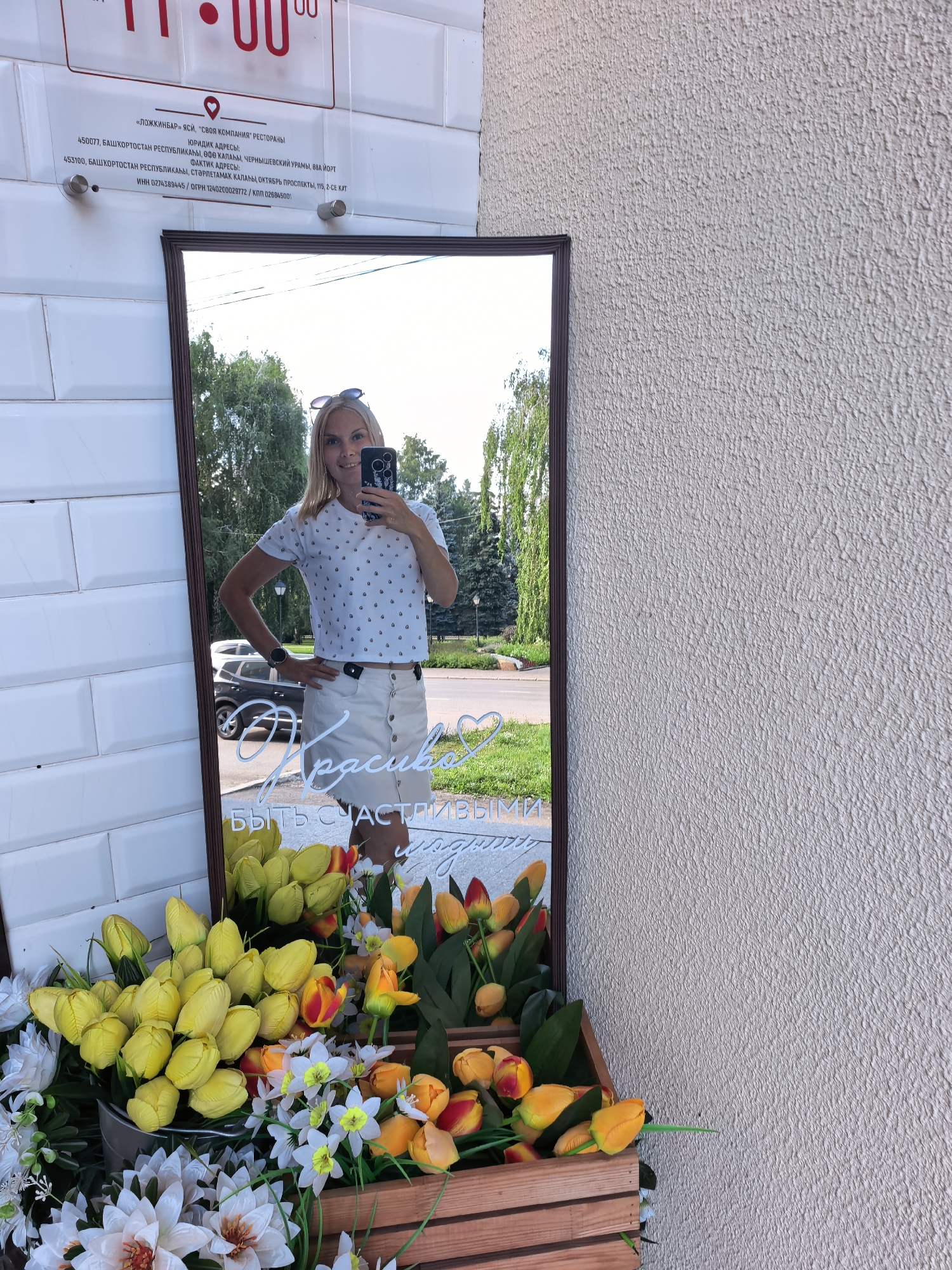 In Sterlitamak, Russia, a blonde woman with a headband is reflected in a mirror, taking a selfie with her phone. She wears a white polka-dot t-shirt and a white skirt. In the foreground, a collection of yellow and red tulips, along with white flowers, are arranged in wooden crates. Behind the woman, a car is visible, and trees line a street. The building's exterior is tiled. A sign above the mirror displays legal and factual addresses for a business.