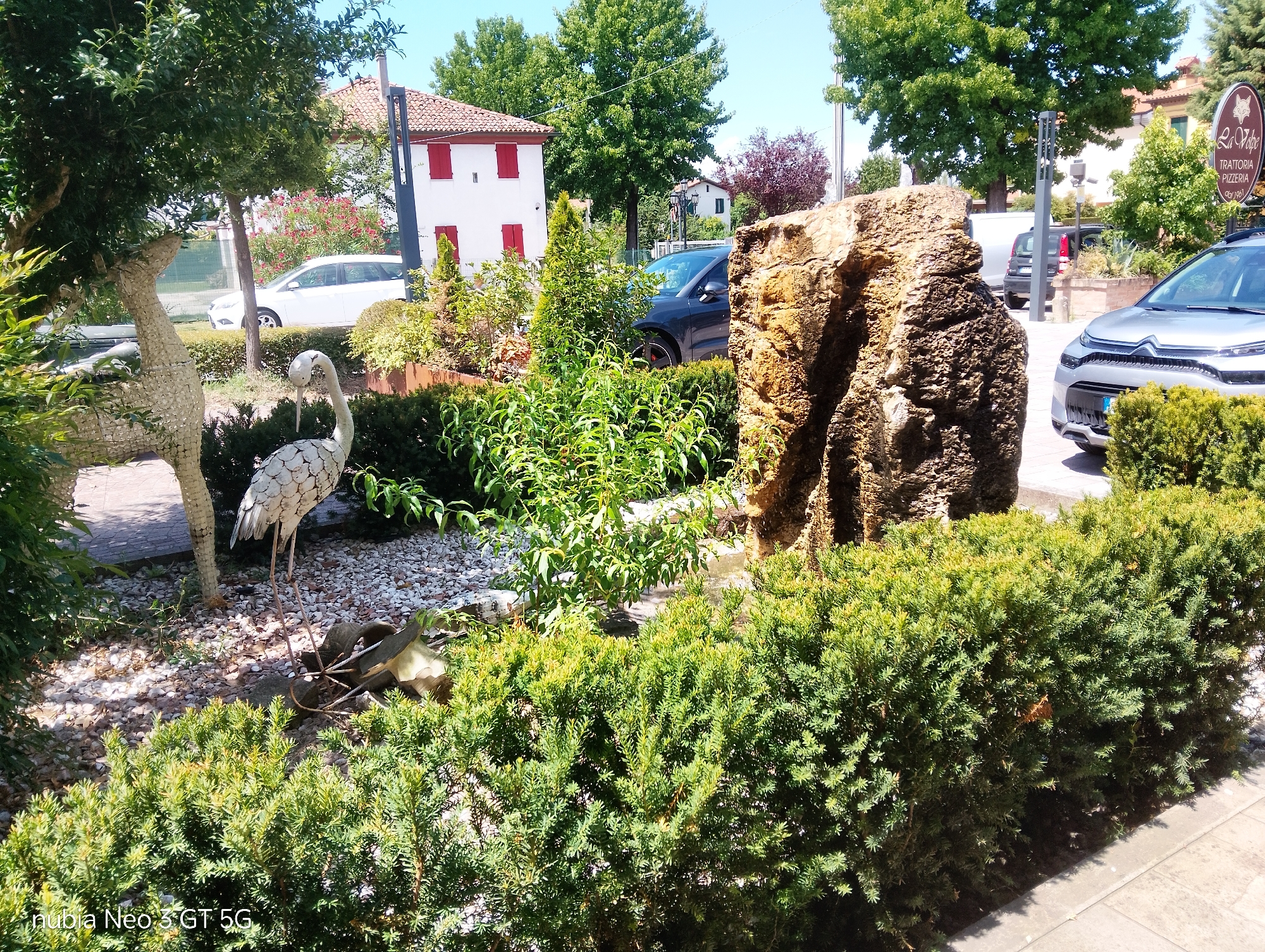The image depicts an outdoor scene in Montegrotto Terme, Italy, likely in front of a restaurant or trattoria. In the foreground, a well-maintained hedge of green shrubs is prominent. Behind the hedge, a large, textured rock formation stands to the right, contrasting with a decorative metal sculpture of a bird on the left. The ground is covered in small white and gray pebbles. Further back, a white building with red-trimmed windows and a terracotta roof is visible, suggesting a traditional Italian architectural style. Several cars, including a silver hatchback and a dark SUV, are parked in the background, indicating a public or commercial area. Lush green trees and foliage surround the scene, contributing to a natural and well-kept environment. A sign for "La Volpe Trattoria Pizzeria" is partially visible on the right, confirming the location as a dining establishment. The bright sunlight and clear sky suggest a daytime setting. There are no people visible in the image.