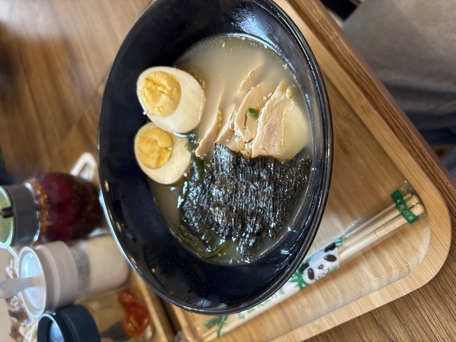 The image displays a close-up, slightly elevated perspective of a meal served on a wooden tray in what appears to be a casual dining establishment in Tbilisi, Georgia.
At the center is a dark, possibly black or dark blue, ceramic bowl filled with a ramen-like dish. The bowl contains a light, opaque broth. To the top right, two halved hard-boiled eggs are present, showcasing their yellow yolks. Below the eggs and to the right, several slices of cooked white meat, identified as chicken, are partially submerged in the broth. A small green garnish, possibly a chopped herb, is visible on one of the chicken pieces. The left side of the bowl is occupied by a substantial portion of dark, crumpled seaweed, likely nori.
The bowl rests on a light-colored wooden serving tray with subtly raised edges. To the left of the bowl, a pair of light-colored wooden chopsticks remains sealed in a clear plastic wrapper. The wrapper features a cartoon panda head design at the top and green text, including what appears to be "开口处" (open here) in Chinese characters, near the bottom.
In the background, positioned on the wooden tray and out of focus, are three condiment bottles. From left to right: a small clear glass bottle with a dark lid containing red chili flakes, a taller clear glass bottle with a white lid containing white granular salt, and another clear glass bottle with a green lid holding dark red chili oil with visible flakes. The surface beneath the tray and the general background is a wooden table with visible grain.