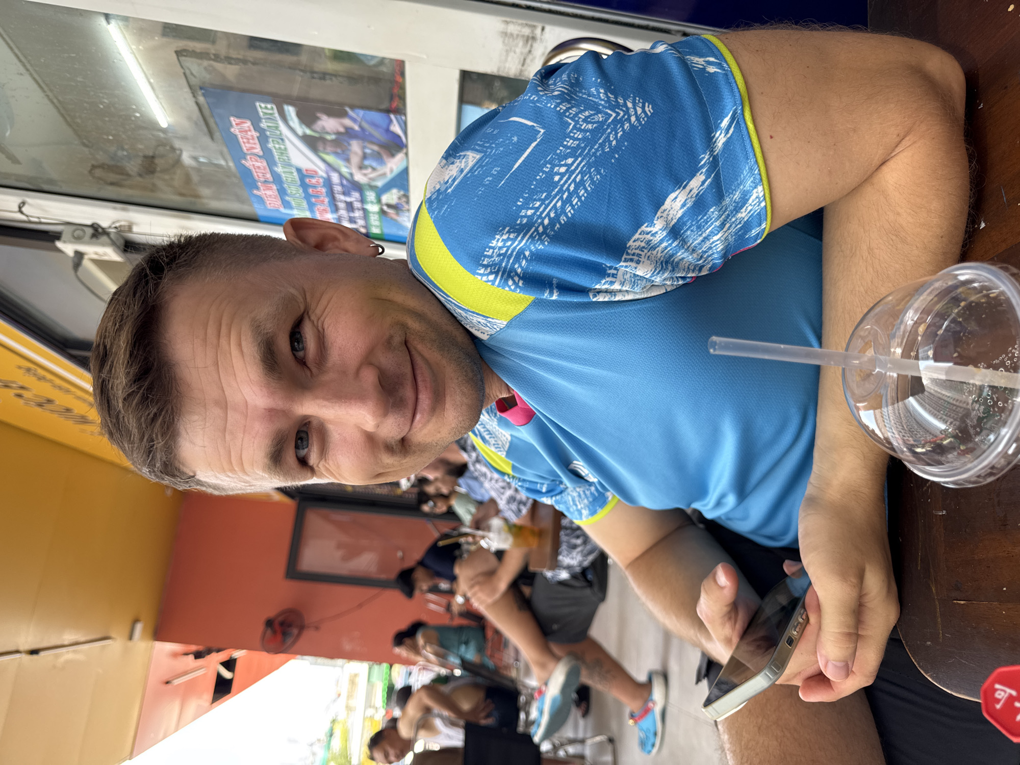 A medium shot depicts a Caucasian male in Bac Nha Trang Ward, Vietnam. He is seated at a wooden table inside a cafe or restaurant, positioned in the foreground. The man is fair-skinned, with short, graying hair, and an earring is visible on his left ear. He is wearing a blue short-sleeved shirt with white and yellow accents. He holds a smartphone in his right hand. Beside him on the table is a clear plastic cup with a straw.
In the blurred background, several other patrons are seated at tables, some holding glasses or drinks. The interior is a mix of orange and yellow tones. A framed picture hangs on the wall behind the patrons. Through an opening, a glimpse of the outside environment is visible.