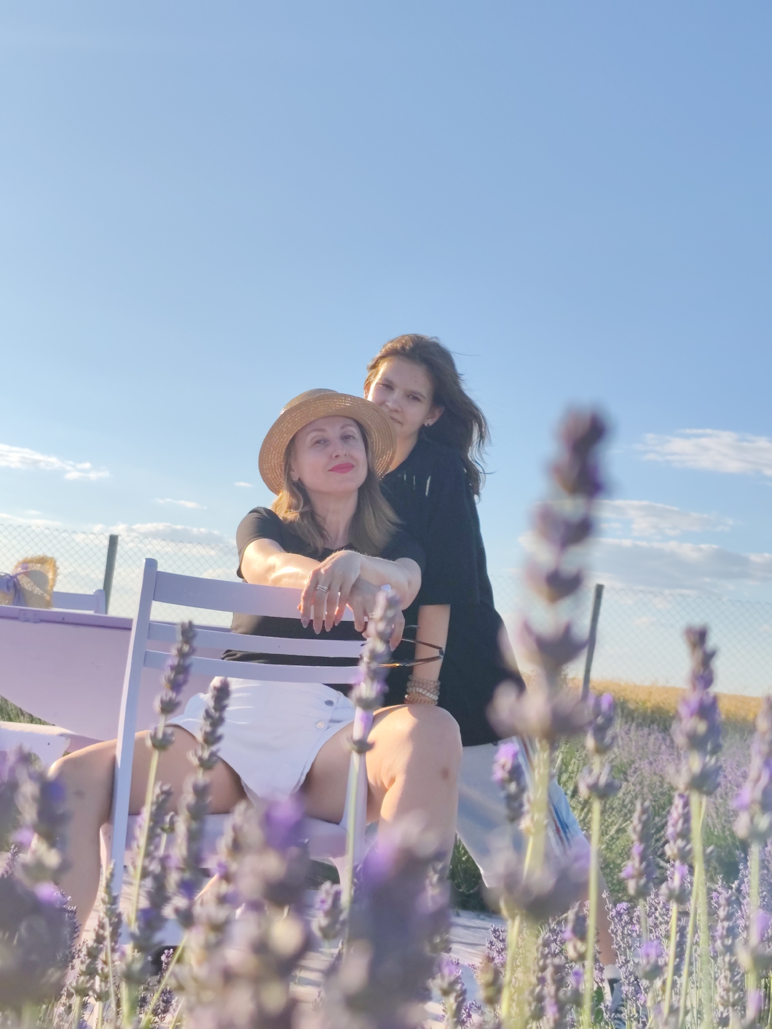 This is a vertical, eye-level shot of two females in a lavender field under a clear blue sky with a few wispy clouds. The sun appears to be setting, casting a warm glow on the scene, suggesting it's late afternoon or early evening. The air seems calm and pleasant, as indicated by the still lavender stalks in the foreground.
In the foreground, a woman in a straw hat and white shorts is seated on a light purple chair, leaning back. She is wearing a black t-shirt and her arms are resting on the chair. Her expression is relaxed, with a hint of a smile. Beside her, a younger female, likely a teenager, stands behind her, also in a black t-shirt. She is looking towards the camera with a gentle smile.
The background features a fence and a field of wheat or dry grass, with more lavender bushes visible. In the far left, a small purple hat with a ribbon is placed on a section of a lilac-colored bench. The overall mood of the image is peaceful and idyllic, capturing a moment of shared enjoyment in a beautiful natural setting. The location is identified as Nechaiane, Ukraine.