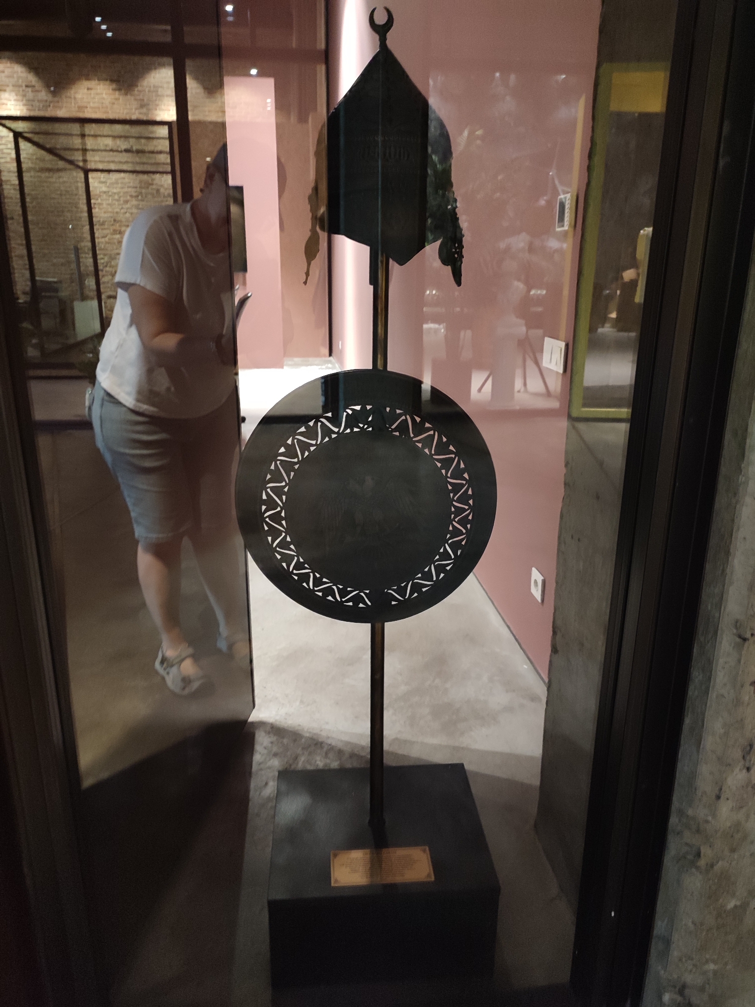 A person wearing a white t-shirt and denim shorts stands in a museum exhibiting a decorative, circular object with an intricate pattern and a crescent moon finial. The object is mounted on a black stand with a small plaque at the base. The scene appears to be within the Koç Museum in Sarıyer, Istanbul, Turkey. The person's reflection is visible in the glass display case.