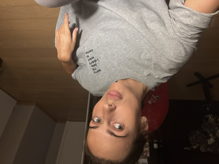 The image captures a light-skinned female individual with light brown hair, pulled back, and green eyes, positioned in an indoor residential environment. She wears a grey short-sleeved t-shirt featuring black text on the left chest that reads: "sorry I'm late. I didn't want to come." Her left arm is visible from the shoulder down, and her right hand rests on her lap. Below the waist, light grey or white trousers are visible. The subject maintains direct eye contact with the camera, exhibiting a neutral facial expression.
The background to the left reveals a dimly lit area with a visible red object, consistent with a chair's backrest, and a dark, slender-legged table holding a vase with dark floral or foliage arrangements. To the right, a light-colored wooden floor extends into a corner, flanked by light-colored walls, indicating an interior room or hallway. The lighting is subdued, with the subject's face more illuminated than the surrounding environment. The scene is an interior space located in Warsaw, Poland. No specific geographical markers or external views indicating a particular district or address within Warsaw are visible in the image.