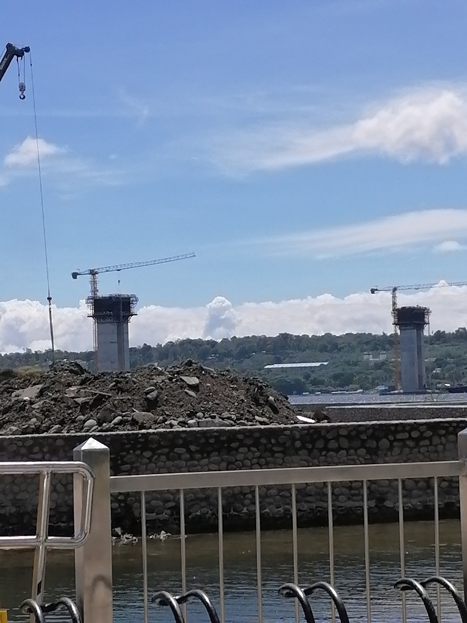 The image depicts a construction site, likely for a bridge, in Davao City, Philippines. Two large concrete pylons are visible, each topped with a construction crane. A third crane is on the left side of the frame. In the foreground, there is a large pile of earth and a low stone wall or embankment. A silver-colored metal railing is also present in the foreground, obscuring a view of the water. Behind the construction site, there is a line of trees or hills, and a long, white building is visible. The sky is blue with scattered white clouds.