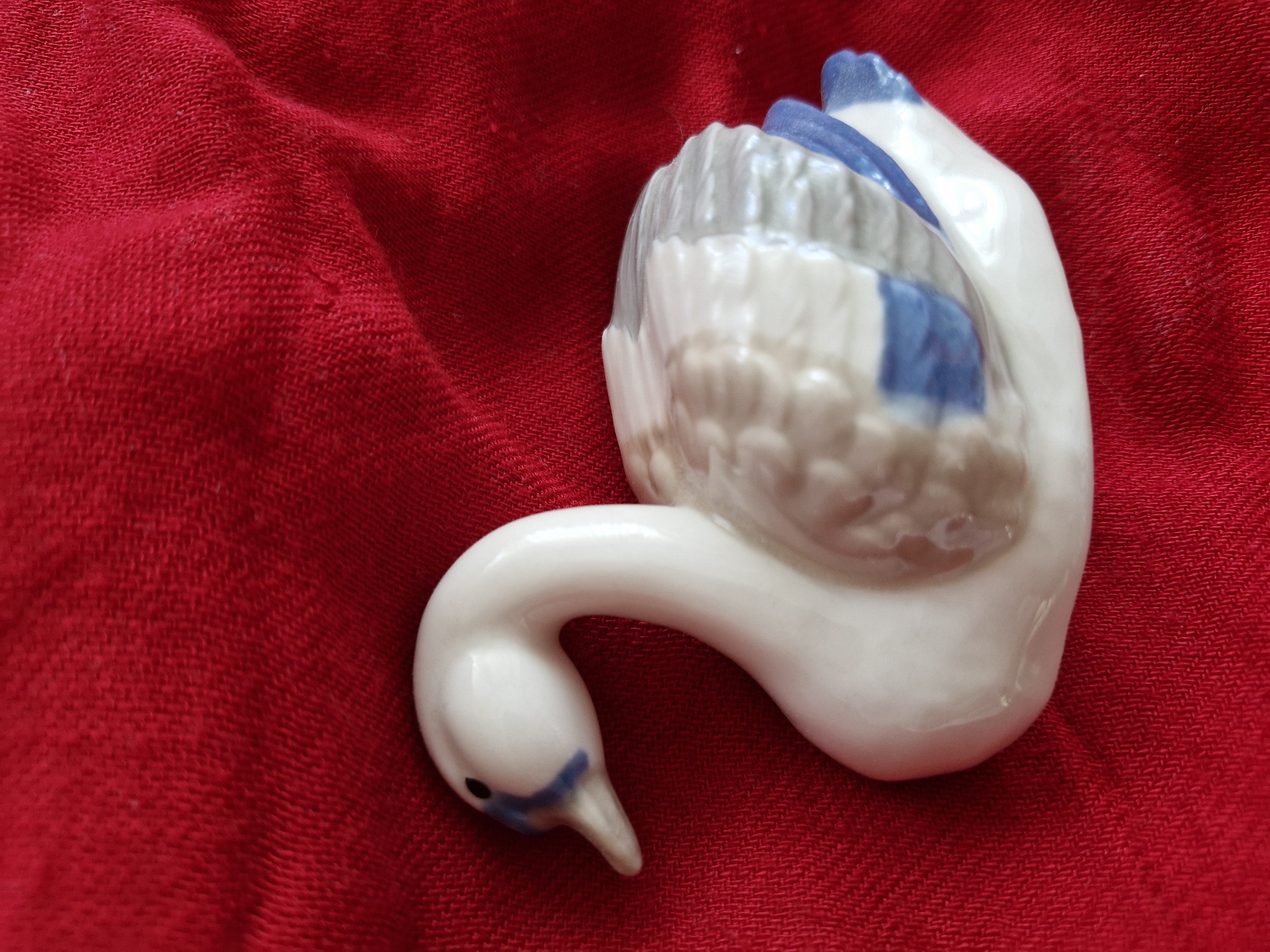 A white porcelain swan figurine with blue accents on its wings and head is placed on a red textured fabric surface. The swan is positioned facing left with its neck curved gracefully upwards. The fabric appears to be a tablecloth or similar textile. The scene is a close-up shot, focusing primarily on the swan and the immediate surrounding fabric. There are no people or any other objects visible in the frame. The lighting suggests an indoor setting. The image does not provide any information about a specific location in Bremen, Germany.