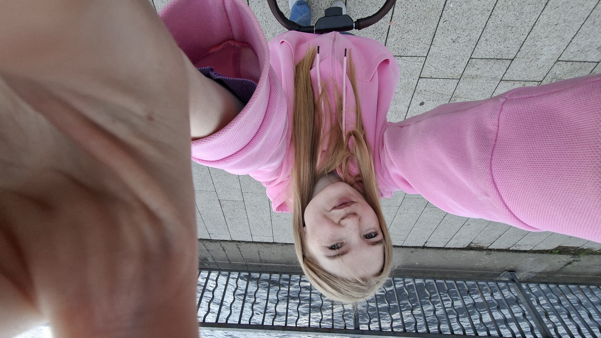 The image is a wide-angle selfie taken in Chaikovsky, Russia, depicting a young adult female outdoors. She has long, straight blonde hair, light-colored eyes, and light freckles across her nose and cheeks. Her lips are lightly pursed, forming a faint, closed-mouth smile as she looks directly into the camera. She is wearing a pink full-zip hooded sweatshirt with long, slightly oversized sleeves and ribbed cuffs; a faint light-colored mark or slight discoloration is visible on the upper left sleeve. Her right hand, holding the camera, partially obscures the upper right corner of the frame. Below her, the dark, curved handle of what appears to be a stroller or pram is visible, along with parts of her blue jeans and white sneakers. The ground is a paved walkway composed of light grey rectangular tiles arranged in a stretcher bond pattern. In the background, a dark-colored metal railing with decorative, wavy vertical bars separates the walkway from a body of water, likely a river or lake, exhibiting a rippled surface.