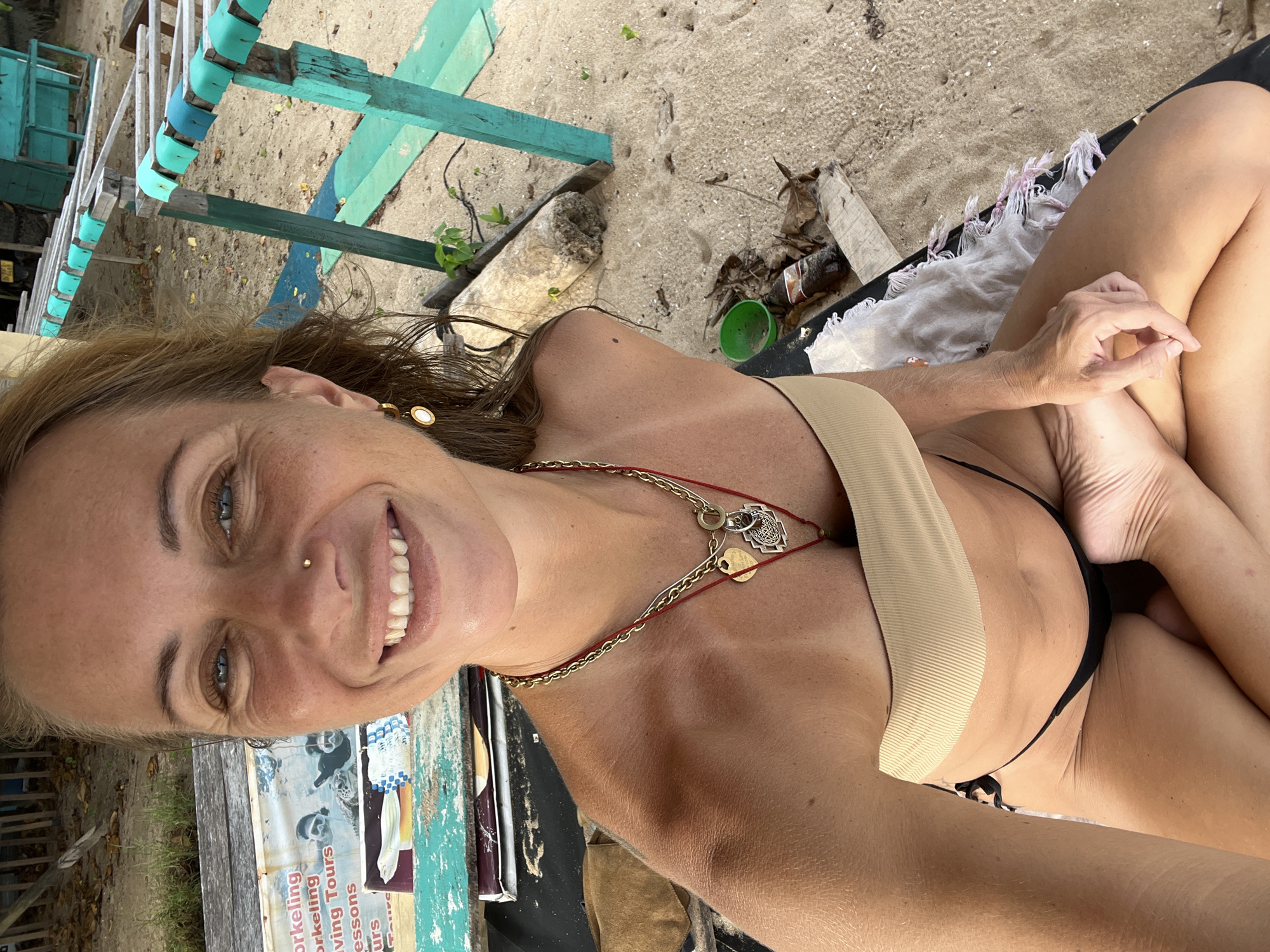 A selfie of a woman in a beige bandeau bikini top and black bikini bottoms taken outdoors on a sandy beach. The woman is sitting cross-legged with her right leg over her left, her right hand resting on her knee in a meditative gesture. She has blonde hair pulled back, blue eyes, and a gold nose ring. She is wearing gold necklaces with charms and gold earrings. In the background, there is a weathered wooden structure with turquoise accents, a piece of driftwood, some dry leaves, and a green plastic cup. There is also a sign behind her with text related to snorkeling and tours. The beach appears to be in Peleana, Sri Lanka, indicated by the context of the image.