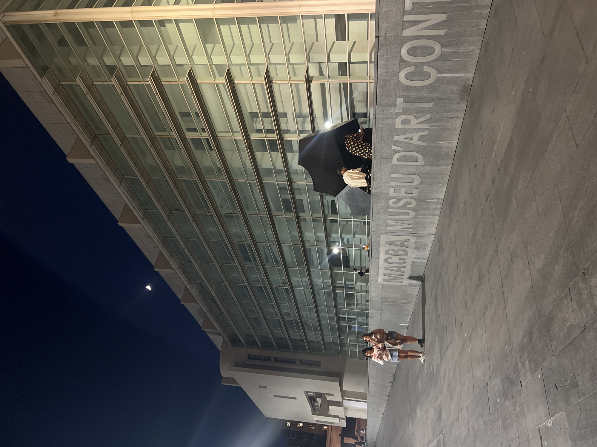 The image shows the Museu d'Art Contemporani de Barcelona (MACBA) at night. The building features a large glass facade and concrete elements. In the foreground, a stone wall with the inscription "MACBA MUSEU D'ART CONT" is visible. Two young women are standing on the pavement in front of the wall, holding drinks. To the right, under a large black umbrella, two individuals are seated. The sky is dark blue with a sliver of the moon visible. The lighting suggests an urban environment at night.