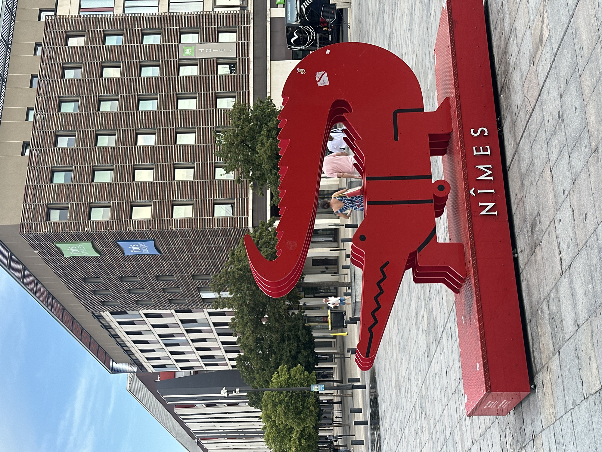 A large, bright red sculpture of a crocodile stands in a city square in Nîmes, France. The sculpture is constructed from flat, angular metal pieces that form the body, head, and tail of the crocodile. The word "NÎMES" is written in white, serif font on the red base of the sculpture. In the background, modern buildings with a grid-like facade are visible, along with trees and paved ground. Several people are walking in the square, and a black bus with "www.hauser-reisen.com" written on its side is parked to the right of the sculpture. The sky is overcast.