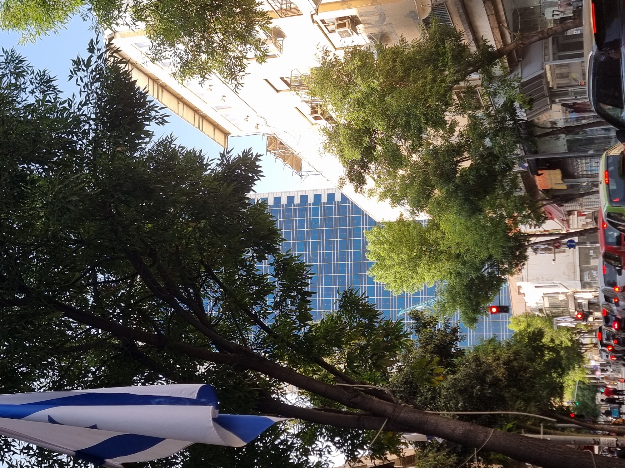 The image captures a street scene in Tel Aviv, Israel, framed by trees and buildings. The viewpoint is tilted upwards, revealing a blend of modern architecture and natural elements. A blue and white striped Israeli flag is prominently displayed in the foreground.
A high-rise building with a blue, grid-like facade stands behind the trees, contrasting with the older, adjacent buildings. These older buildings feature a mix of residential and commercial elements, including balconies and window air conditioning units.
A busy street runs along the side of the image, filled with cars stopped at a traffic light, which is visible as a red signal. Pedestrians can be seen walking along the sidewalks. The overall environment suggests a lively urban setting with a mix of architectural styles and vibrant street activity.