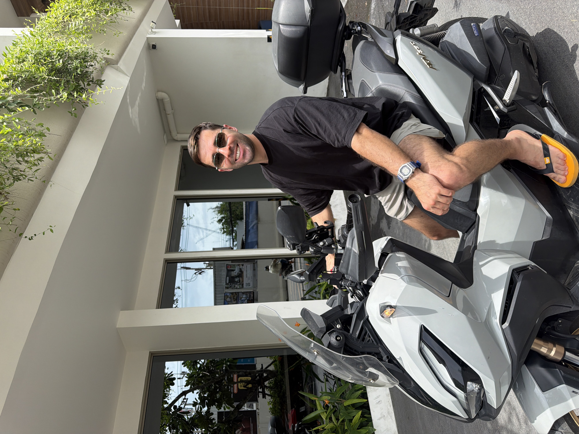 A man is sitting on a white and gray Can-Am motorcycle. He is wearing a black t-shirt, khaki shorts, sunglasses, and a blue wristwatch. He has one leg folded across the seat, and is wearing a yellow and black sandal on that foot. The scene is outside of a building with large windows. The location appears to be Choeng Thale, Thailand, based on visual context. Green plants are growing on the exterior of the building. The sky is visible through the windows.