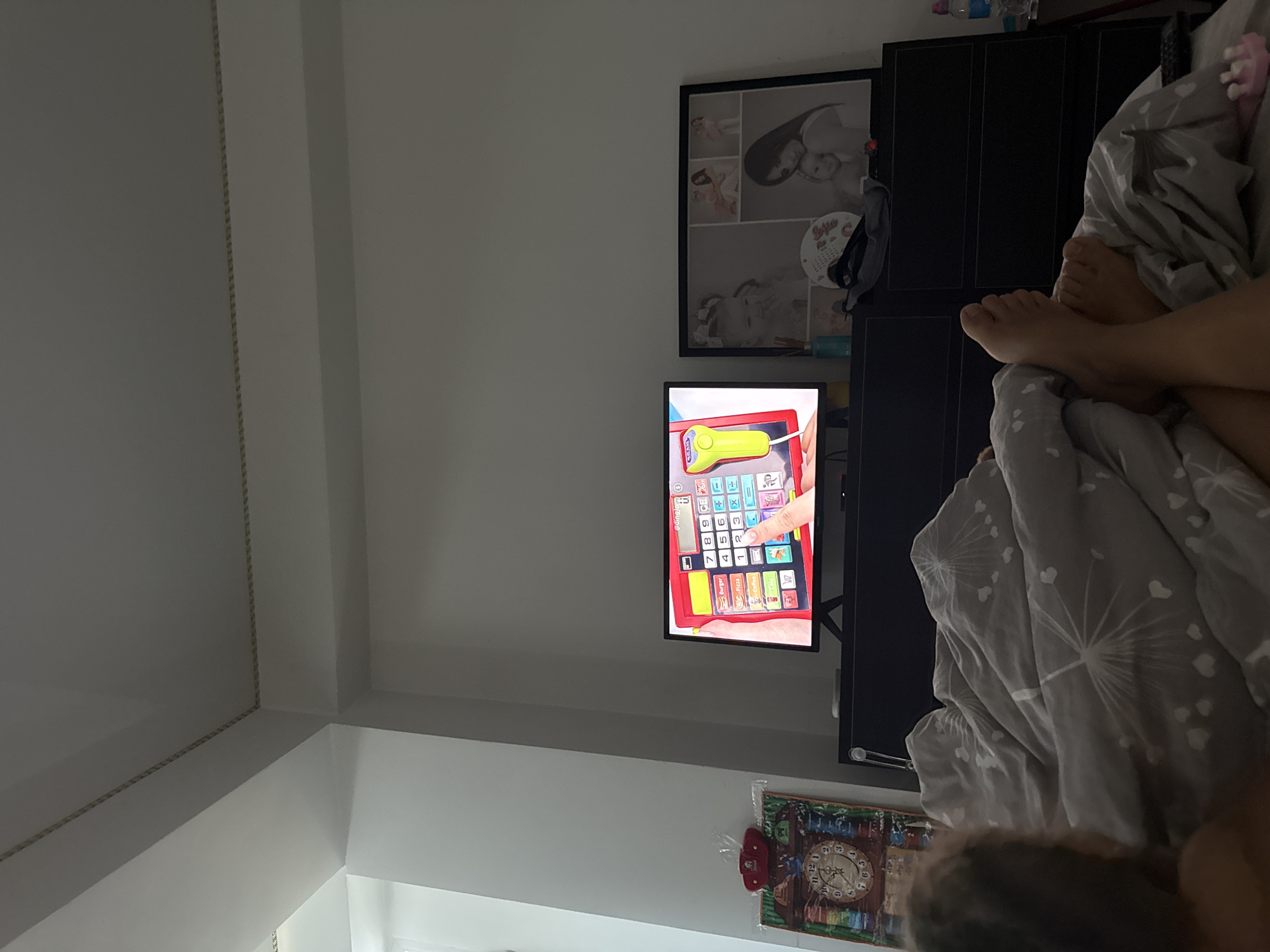 This is a low-angle, indoor shot of a bedroom in Chisinau, Moldova, focusing on a person lying in bed watching television. The person, partially visible from the back and legs, is under a grey duvet with a dandelion pattern. The television screen displays a game or application with a cash register interface, featuring buttons for food items like "Burger," "Pizza," "Coffee," and "Cola," along with a numeric keypad and a scanning device. To the left of the television, on a black dresser, is a decorative item resembling a clock face and a small red object. To the right of the television, also on the dresser, is a framed collage of photos featuring women and children, and a small blue vase with a sprig of greenery. The room appears to be dimly lit, with light coming from the television screen and potentially a window out of frame. The ceiling has decorative molding.