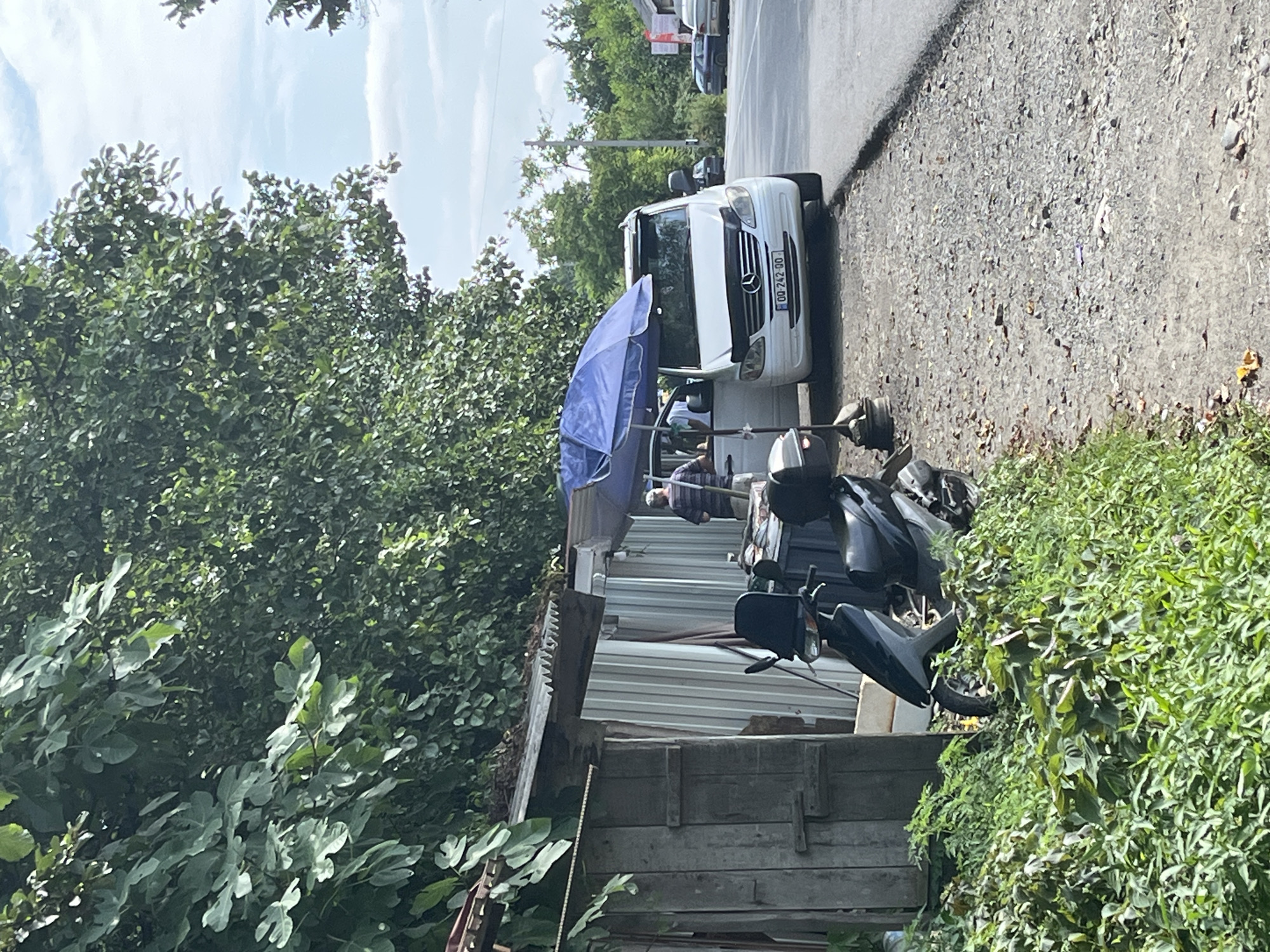 On a sunny day in the city of Ghrmaghele's Temi, Georgia, a white Mercedes-Benz Vito van is parked on the side of a road. The van's passenger door is open, and a person wearing a striped shirt stands near it, holding a pole. A blue umbrella is positioned above the van, providing shade. In front of the van, a black scooter is parked. To the left, there is a weathered wooden structure and a corrugated metal shed. Lush green foliage surrounds the scene, with trees visible in the background under a partly cloudy sky. The road appears to be asphalt, with a gravel shoulder. The license plate on the van reads "QQ-242-QQ".