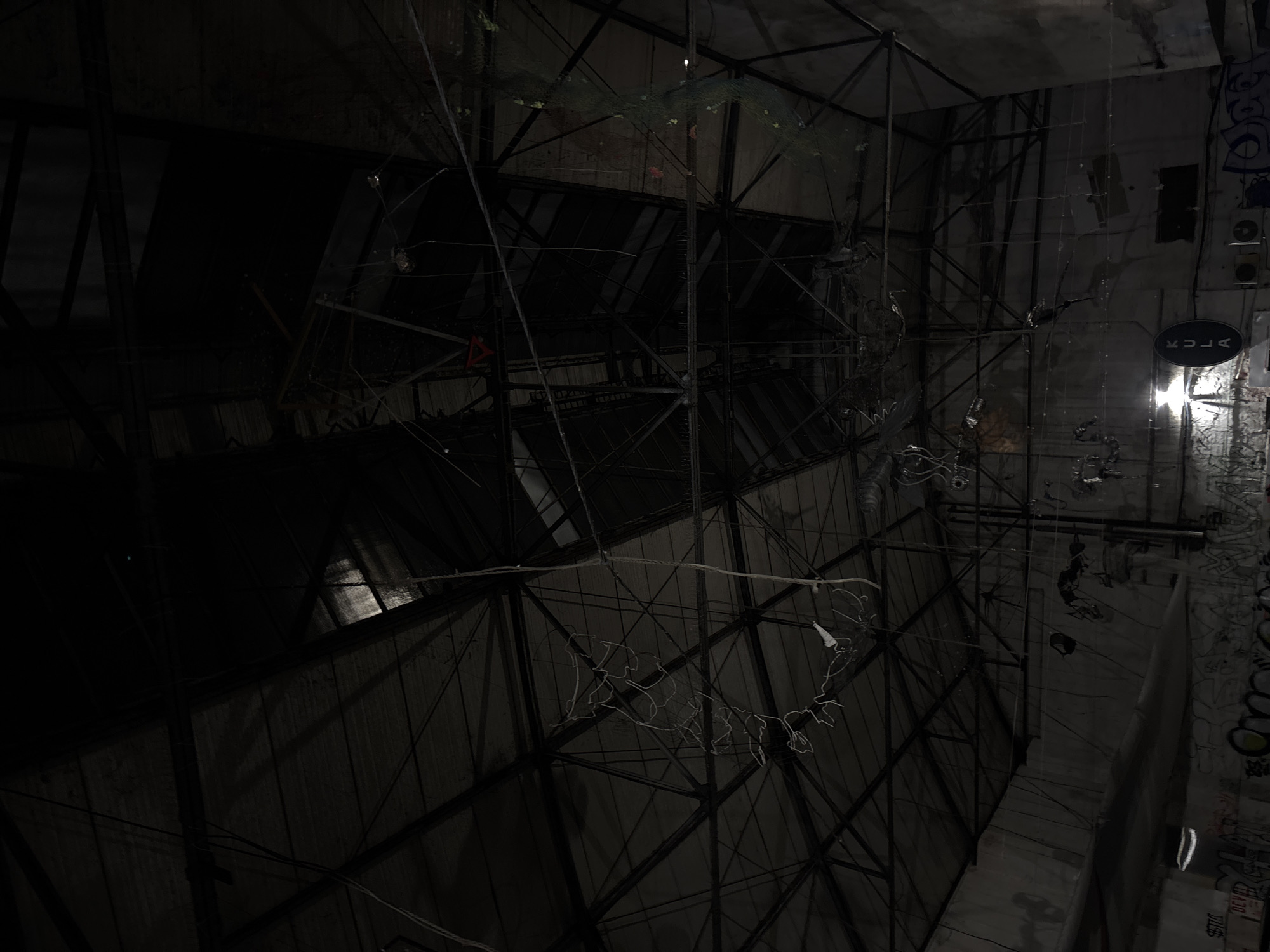 The image depicts an interior view of an abandoned structure. The structural framework is composed of metal beams, creating a grid-like pattern. Various wires and hanging objects are visible. Graffiti is present on the walls in the brighter area. The illumination is uneven, casting significant portions of the structure into darkness.
The location is Belgrade, Serbia.