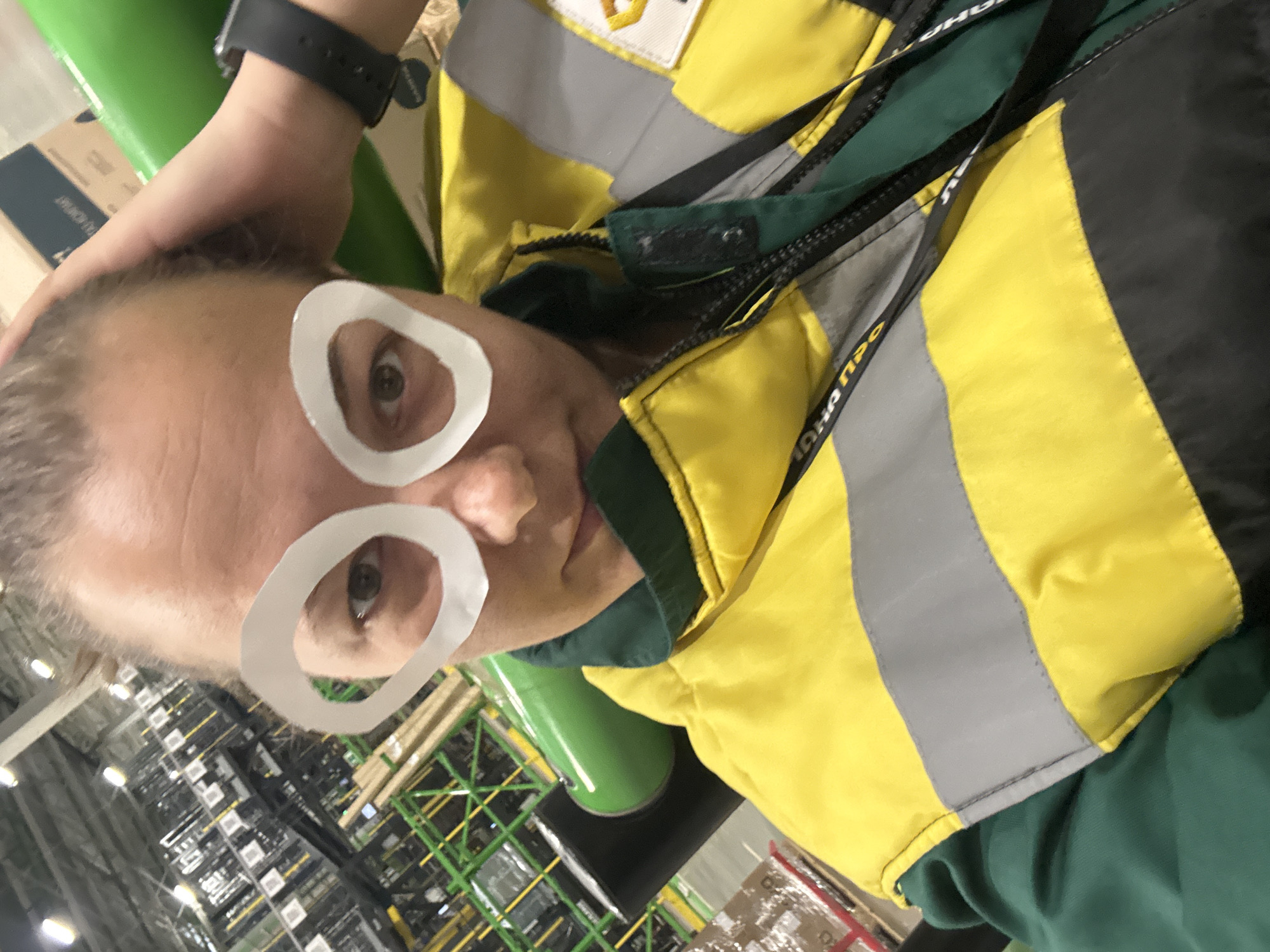 A person is posing with white paper circles over their eyes, resembling glasses. The person has fair skin, short hair, and a dark smartwatch on their left wrist. They are wearing a green and yellow safety jacket with reflective gray stripes. The background shows warehouse shelving filled with boxes and materials, suggesting an industrial or storage facility. The location appears to be indoors, in Lupanovo, Russia.
