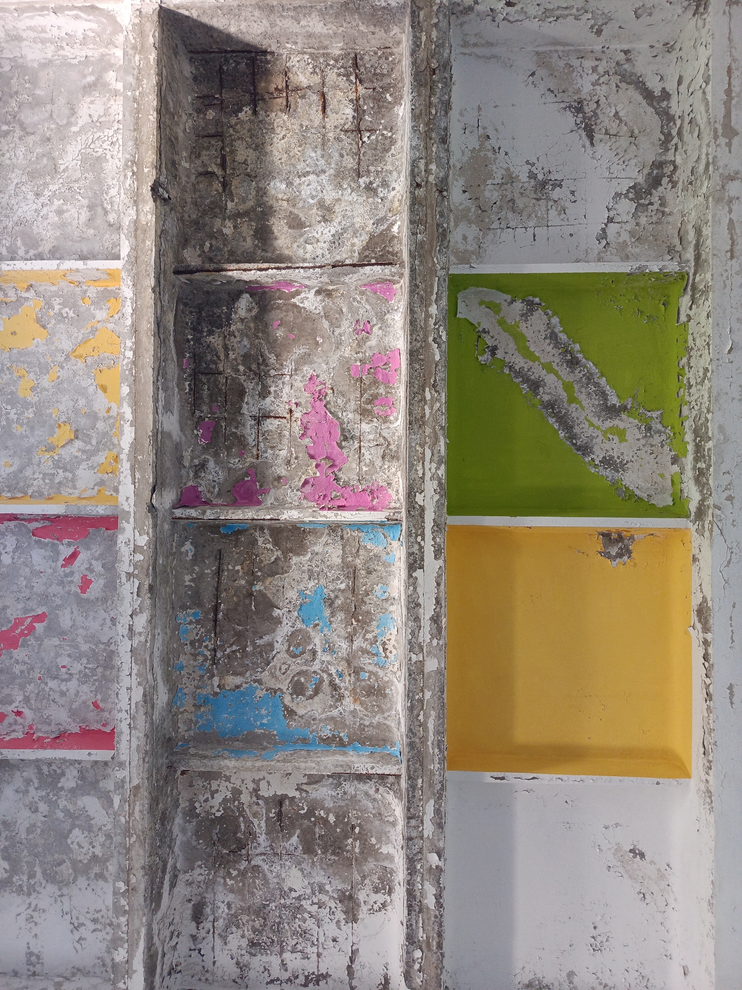This is a close-up, top-down view of a weathered concrete structure with sections of peeling paint in various colors. The structure appears to be part of an outdoor architectural element, possibly a ceiling or a wall, exhibiting signs of decay and age. The concrete is rough and textured, with visible rebar in some areas, indicating it may be under construction or in a state of disrepair.
On the left side, vertical concrete columns or sections are separated by a central channel. These left sections have patches of flaking white paint over grey concrete, with horizontal bands of color. The top band visible is yellow, followed by a pink band, and then another pink band.
The central channel is a darker, rougher concrete with exposed rebar and shadows. It also shows patches of peeling paint, with glimpses of pink and blue underneath.
On the right side, there are larger, smoother rectangular sections. The top section is a vibrant green with streaks of grey and white, suggesting the paint is also peeling or worn. Below the green section is a solid yellow rectangle. The rightmost edge of the image shows a light grey surface with extensive peeling paint.
The lighting suggests an outdoor setting, with a strong shadow cast from the upper left into the central channel. The overall impression is one of urban decay, with a stark contrast between the rough, aged concrete and the remaining bright, yet damaged, paintwork. This image was taken in Baku, Azerbaijan.