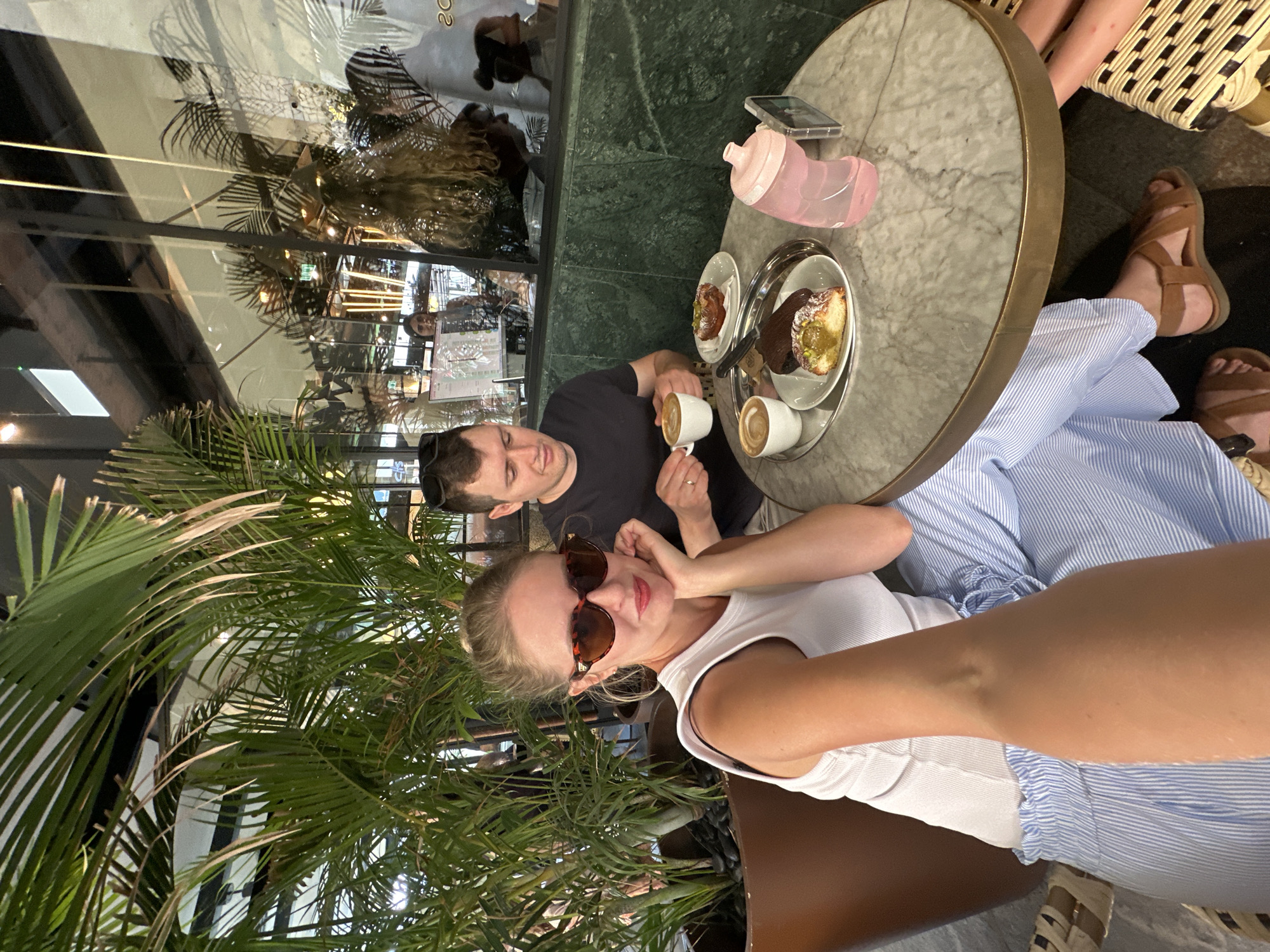A medium shot captures a couple seated at a marble-topped table in a cafe, possibly located in Herzliya, Israel. The woman, in the foreground and to the left, wears a white tank top and blue and white striped pants, accessorized with large, tortoiseshell sunglasses and her hand resting on her chin. The man, to her right, is dressed in a dark t-shirt and holds a cup of coffee. On the table between them are two plates with pastries, two more cups of coffee, a pink water bottle, and a cell phone. Behind the couple, lush green foliage from a large potted plant is visible. The background features a reflective surface, likely a window or glass wall, showcasing a glimpse of the cafe's interior with patrons and decor.