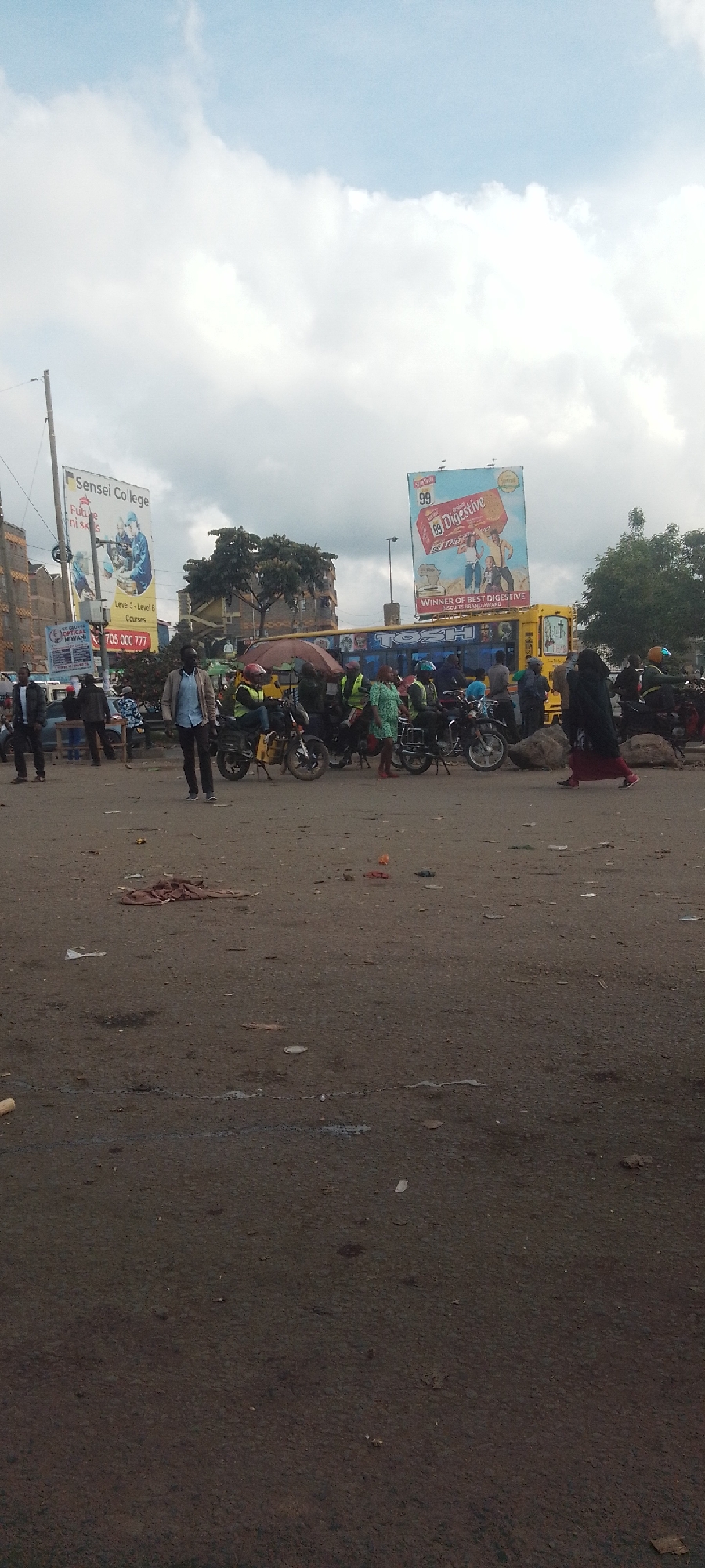 This is a daytime scene in Nairobi, Kenya, set in what appears to be a busy intersection or open area. The ground is uneven and unpaved with visible debris scattered across it. In the background, there is a mix of structures, including buildings and billboards, advertising "Sensei College" and "99 Digestive" biscuits. A yellow bus with "TOSH" written on its front sits amid motorcycles and people. Several people are standing or walking in the vicinity, including men in casual wear, one woman wearing a dark head covering. The overall atmosphere suggests a crowded and active urban environment.
