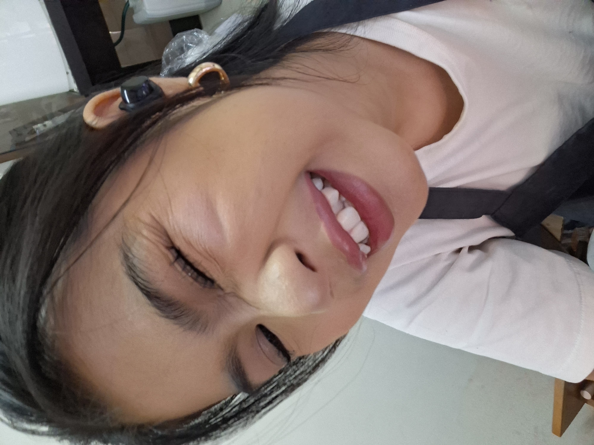 A close-up, low-angle selfie shows a young woman with dark hair and fair skin, squinting her eyes and smiling. She is wearing a white t-shirt and a black strap across her shoulder, possibly part of an apron. A black earbud is visible in her right ear, and a gold hoop earring adorns her left ear. Her lips are painted a bright pink. The background is out of focus, but suggests an indoor environment with a hint of a white wall and some furniture. No specific location in Kuta, Indonesia is identifiable from the image.