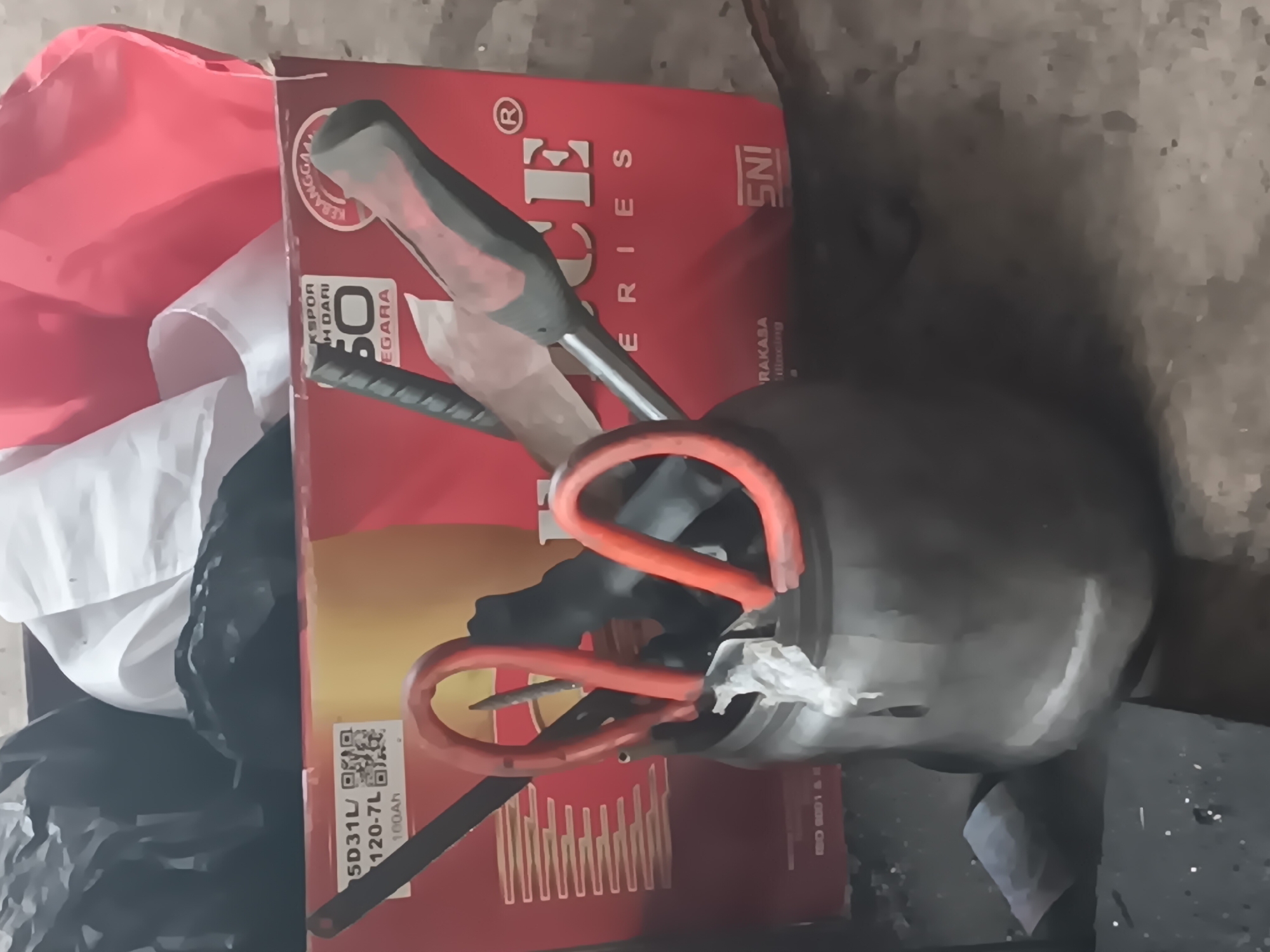 This image captures a cluttered scene likely within a workshop or storage area in Tlogo, Indonesia. In the foreground, a dark, metallic container holds various tools, most notably a pair of large red-handled scissors. A screwdriver with a red and black handle is also visible, positioned behind the scissors and pointing upwards. Behind this container, a large red cardboard box is prominent. It features yellow and white branding with text that includes "EKSPOR", "H DARI", "50", "EGARA", and "SERIES", along with a QR code. The box appears to contain black and white plastic bags, suggesting storage or packaging materials. The ground is a rough, concrete-like surface. The overall impression is one of organized disarray, typical of a functional workspace.