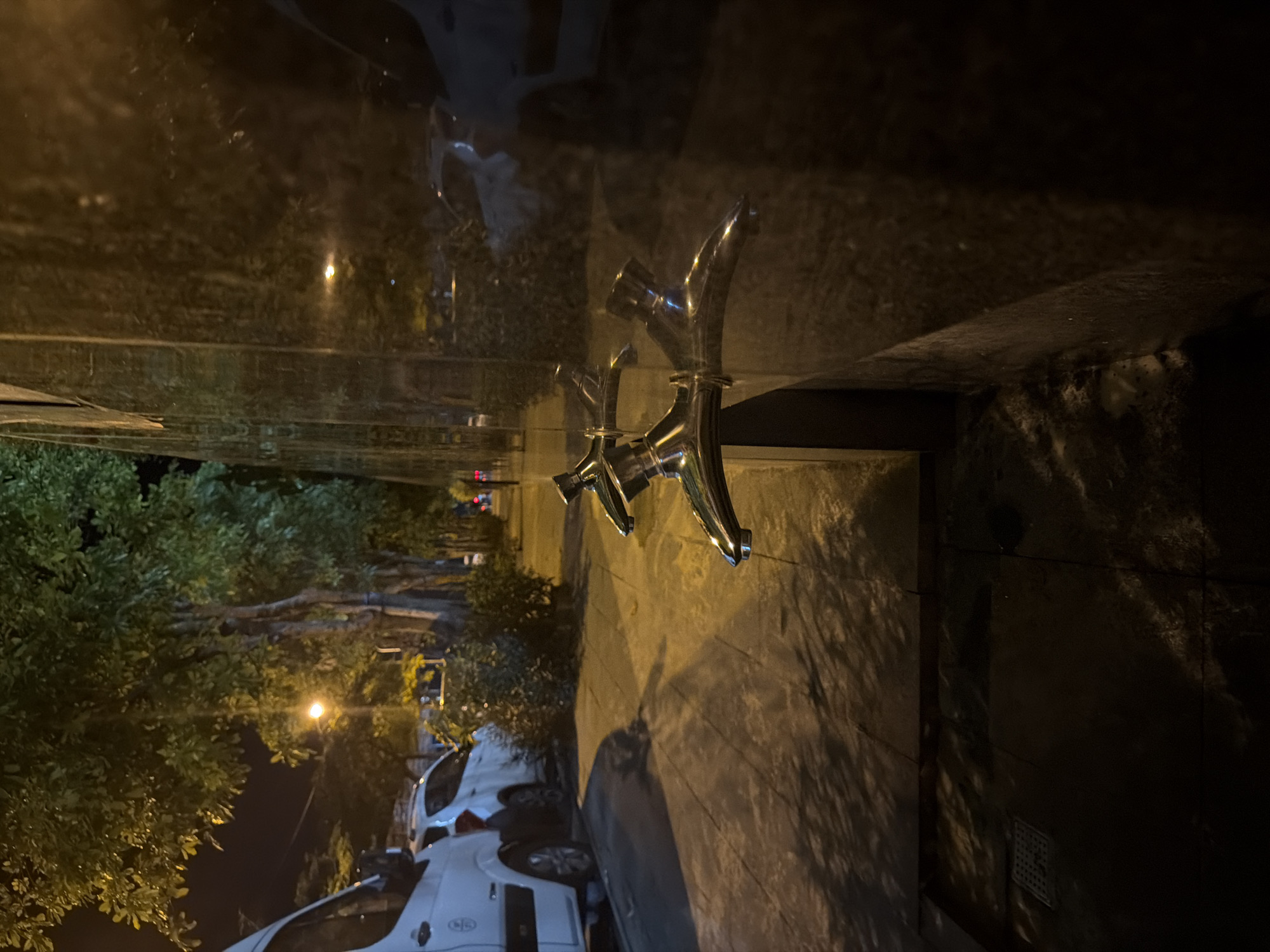 The image depicts a nighttime scene on a street in Bac Nha Trang Ward, Vietnam. In the foreground, two chrome water faucets are visible, mounted on a dark, reflective surface, possibly a wall or pillar. Their polished surfaces reflect the surrounding light. To the left, a sidewalk runs parallel to the street, with a light-colored van and a white car parked along the curb. Trees line the sidewalk, their foliage illuminated by a streetlamp or other light source, casting dappled shadows on the ground. The environment appears urban, with elements of street infrastructure and parked vehicles. The overall lighting suggests it is after dark, with artificial lights providing the primary illumination.