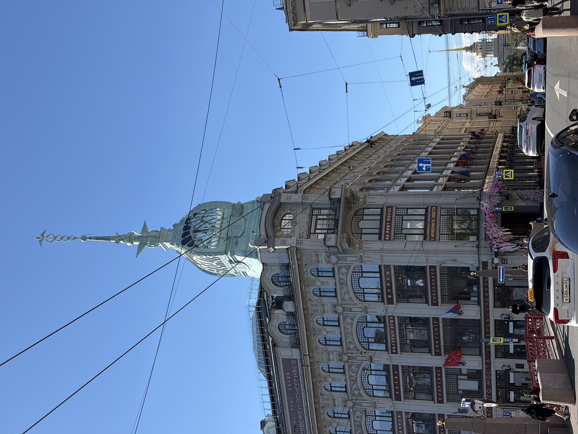 This is a street-level, eye-level, medium-long shot of the Singer House building in Saint Petersburg, Russia, on a clear, sunny day. The iconic Singer House, also known as the Dom Knigi (House of Books), is a prominent landmark located on Nevsky Prospekt. The building is characterized by its elaborate Belle Époque architecture and a distinctive glass dome topped with a stylized eagle sculpture.
In the foreground, a street scene unfolds with several vehicles, including a white Haval SUV with a license plate starting with B586OM, and other cars moving along the street. Pedestrians are visible walking on the sidewalks and crossing the street. The street is lined with buildings, and overhead, a complex network of electrical wires crisscrosses the sky, a common sight in the city.
The Singer House itself occupies the left and center portions of the frame. Its facade displays multiple stories with numerous windows, adorned with ornate detailing. Banners and flags hang from the building, some displaying foreign language text, likely indicating retail establishments within. Pink floral decorations cascade down from a balcony area on the building, adding a decorative touch.
Further down the street, in the background, the spire of the Admiralty Building can be seen piercing the clear blue sky, a common visual reference point in Saint Petersburg's cityscape. Traffic signs, including directional arrows and pedestrian crossing indicators, are strategically placed along the roadway. The overall scene depicts a bustling urban environment with historical architecture and everyday city life.