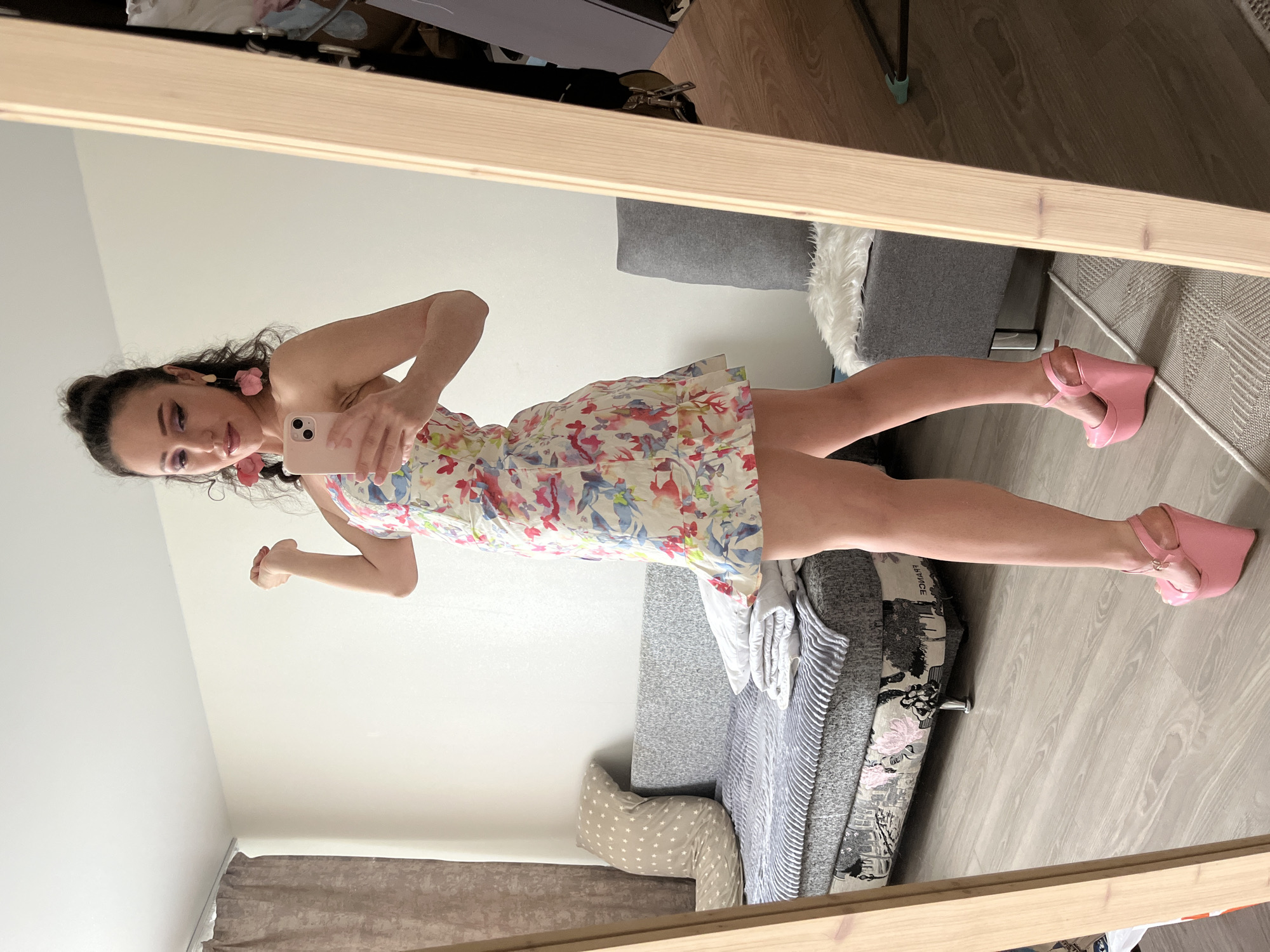 A woman is taking a mirror selfie in a room. She is wearing a floral print mini dress and pink wedge heels. She has her hair up in a bun and is wearing large pink earrings. In the background, there is a sofa with pillows and a draped blanket, a grey armchair with a white rug, and a mirror with a wooden frame. The floor is light-colored wood. The scene appears to be an indoor residential setting. There is no indication of this location being in the city of Kazan, Russia.